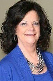 A woman wearing a blue jacket and a pearl necklace is smiling for the camera.