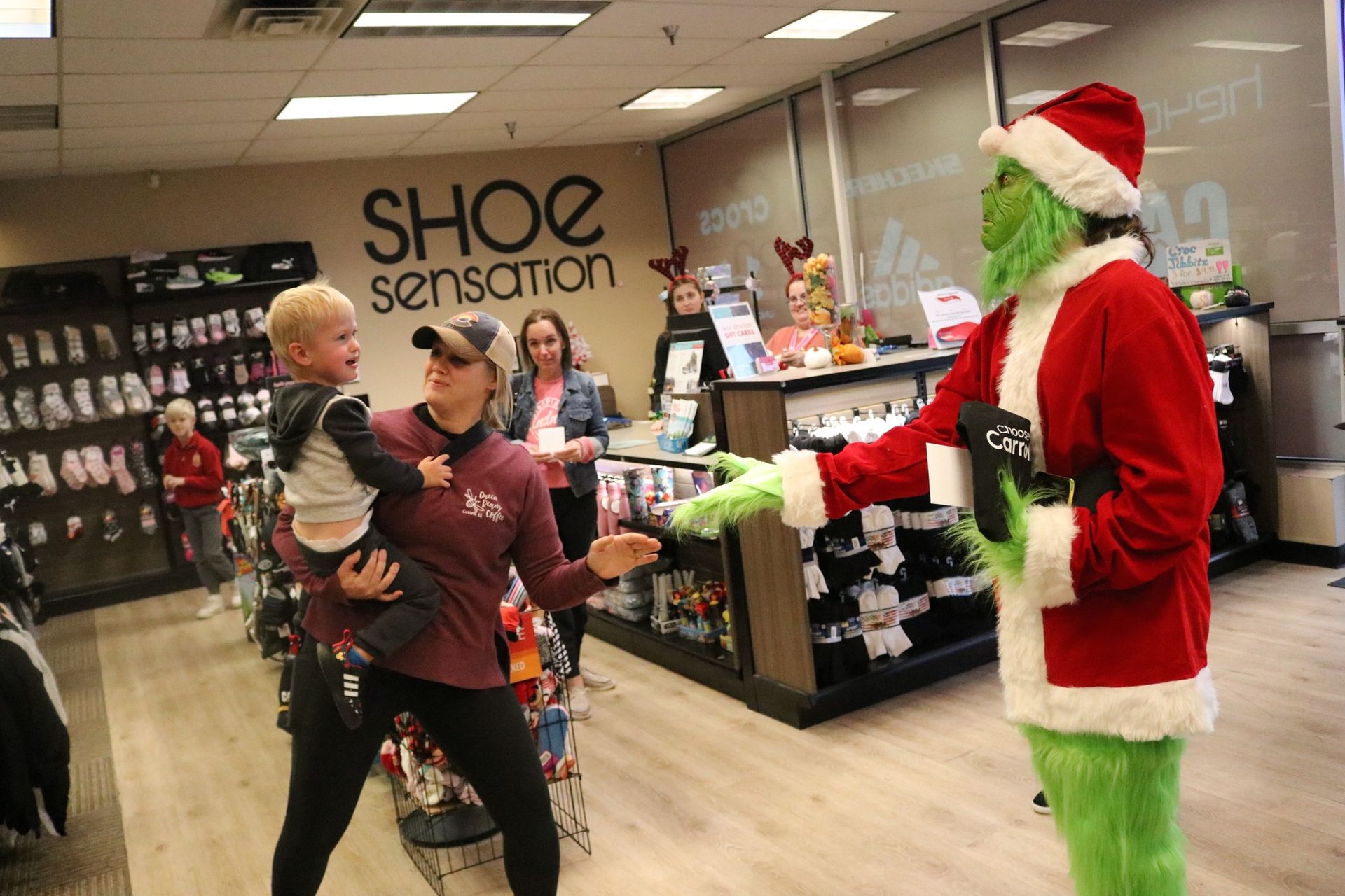 A woman is holding a baby and talking to a grinch in a shoe store.