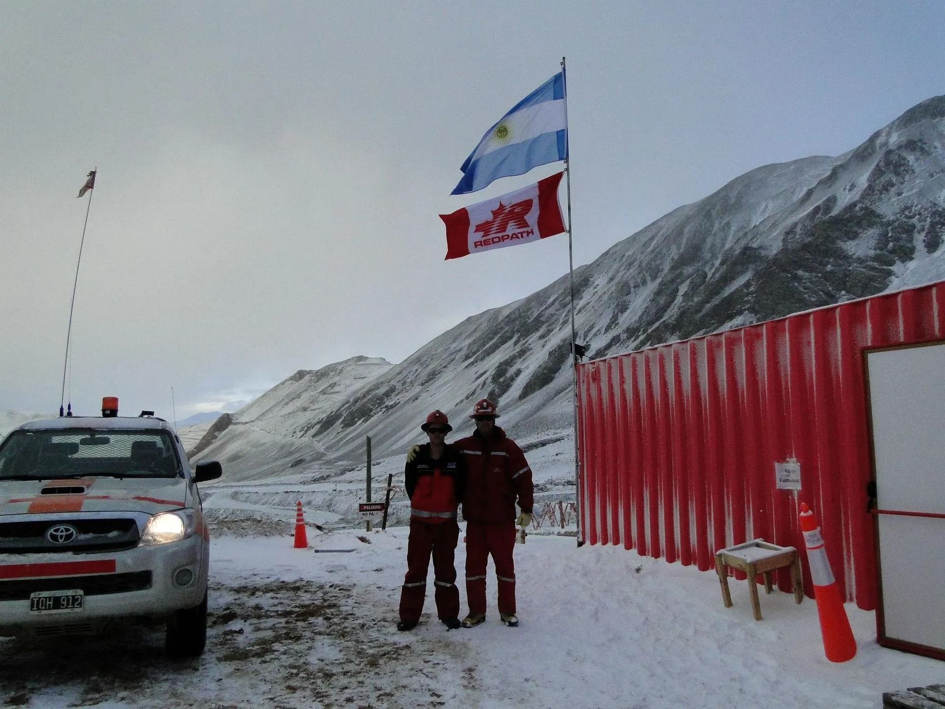 Dos trabajadores junto a un puesto de control nevado en la carretera, con banderas, un camión y vallas rojas en un paso de montaña.