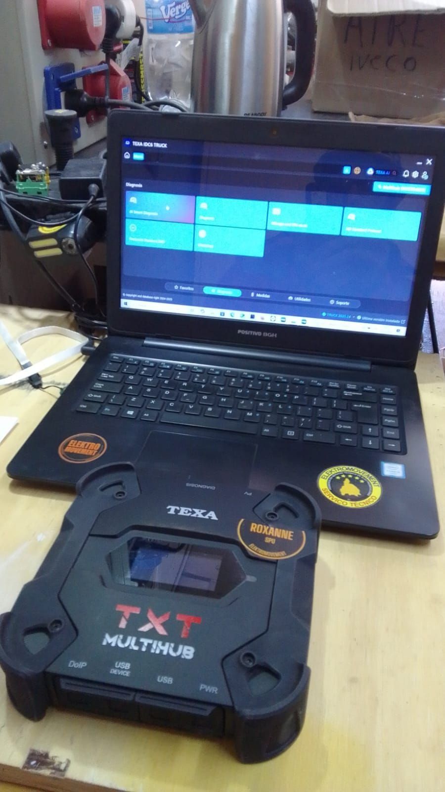 Sobre el escritorio hay un ordenador portátil que muestra un programa informático, con un dispositivo TXT MultiBus negro delante y cables a su alrededor.