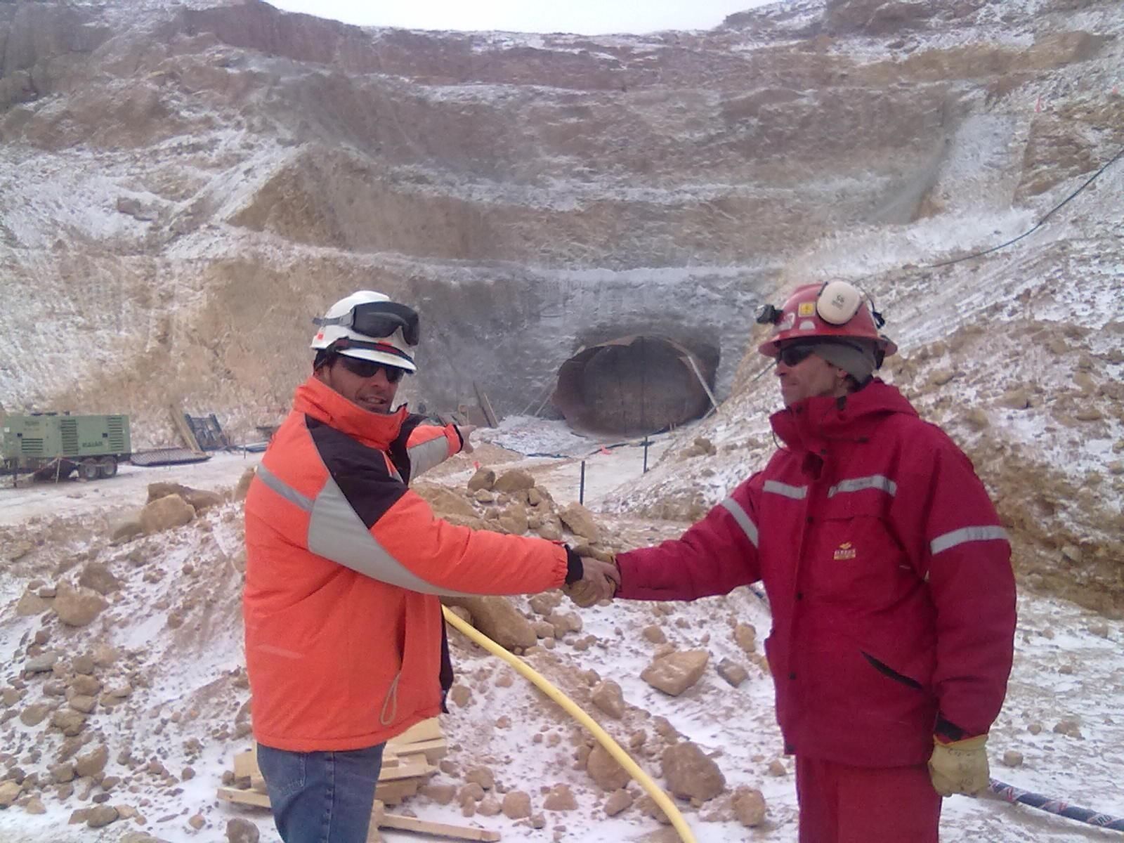 Dos trabajadores con ropa de invierno se dan la mano en la entrada de una mina nevada.