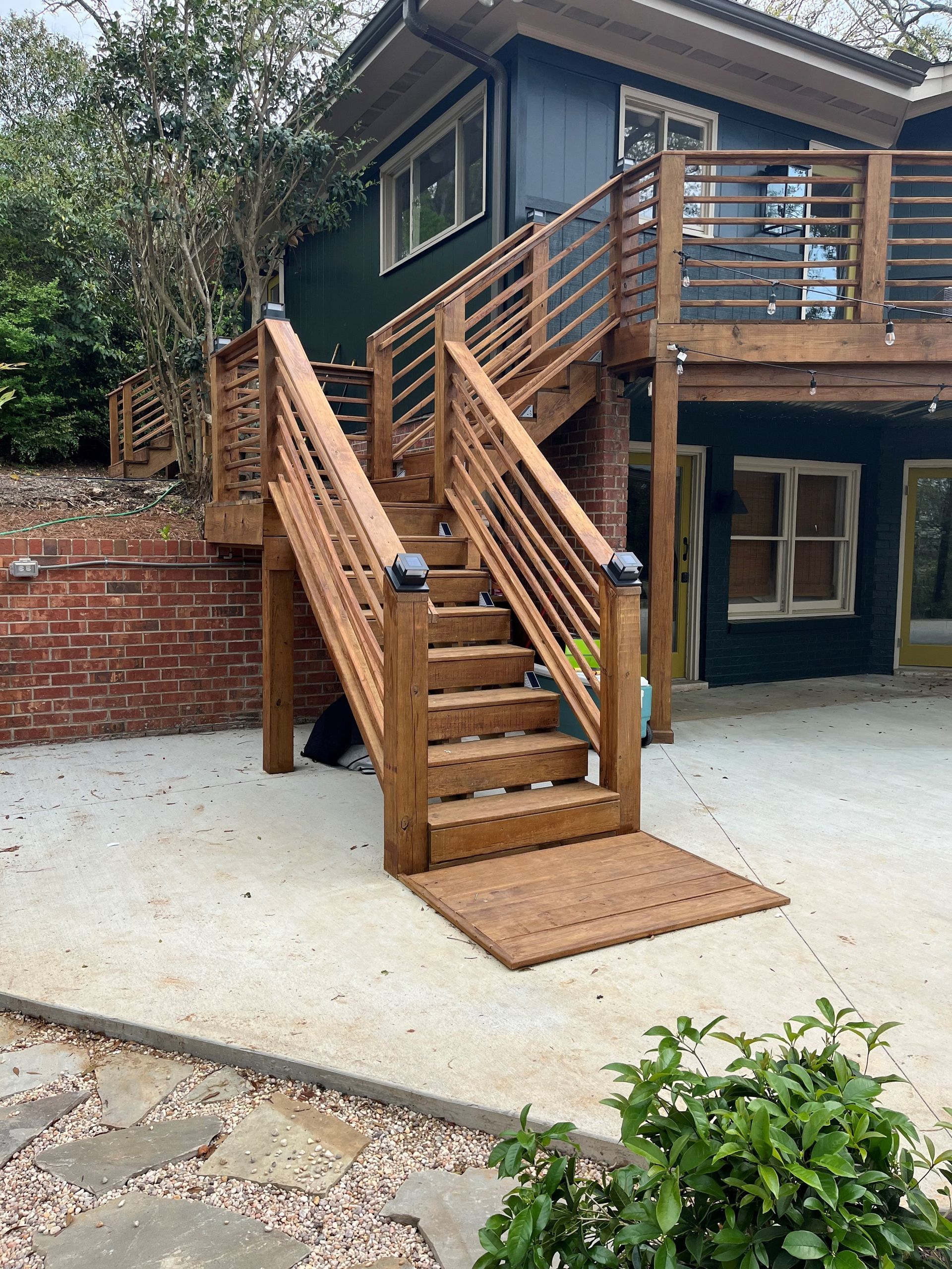 Wooden outdoor staircase and deck attached to a dark house, with brick wall and concrete patio.
