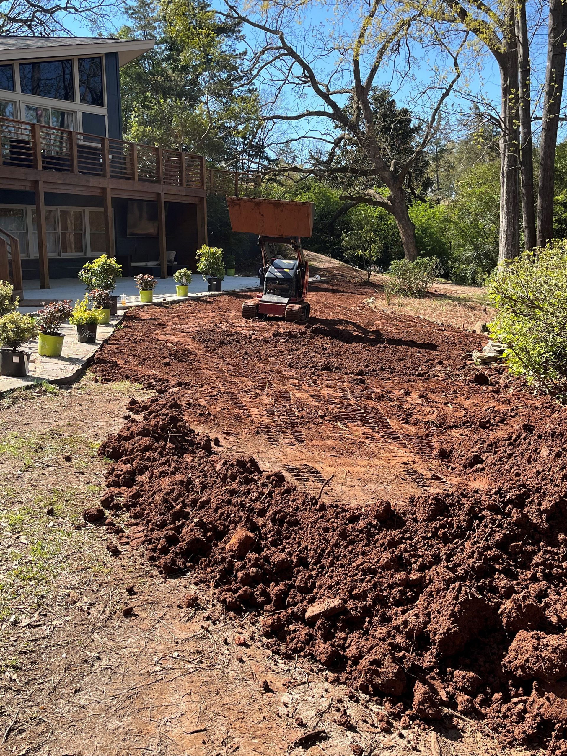 Garden bed with fresh mulch beside a house, with trees and a wheelbarrow in the background.