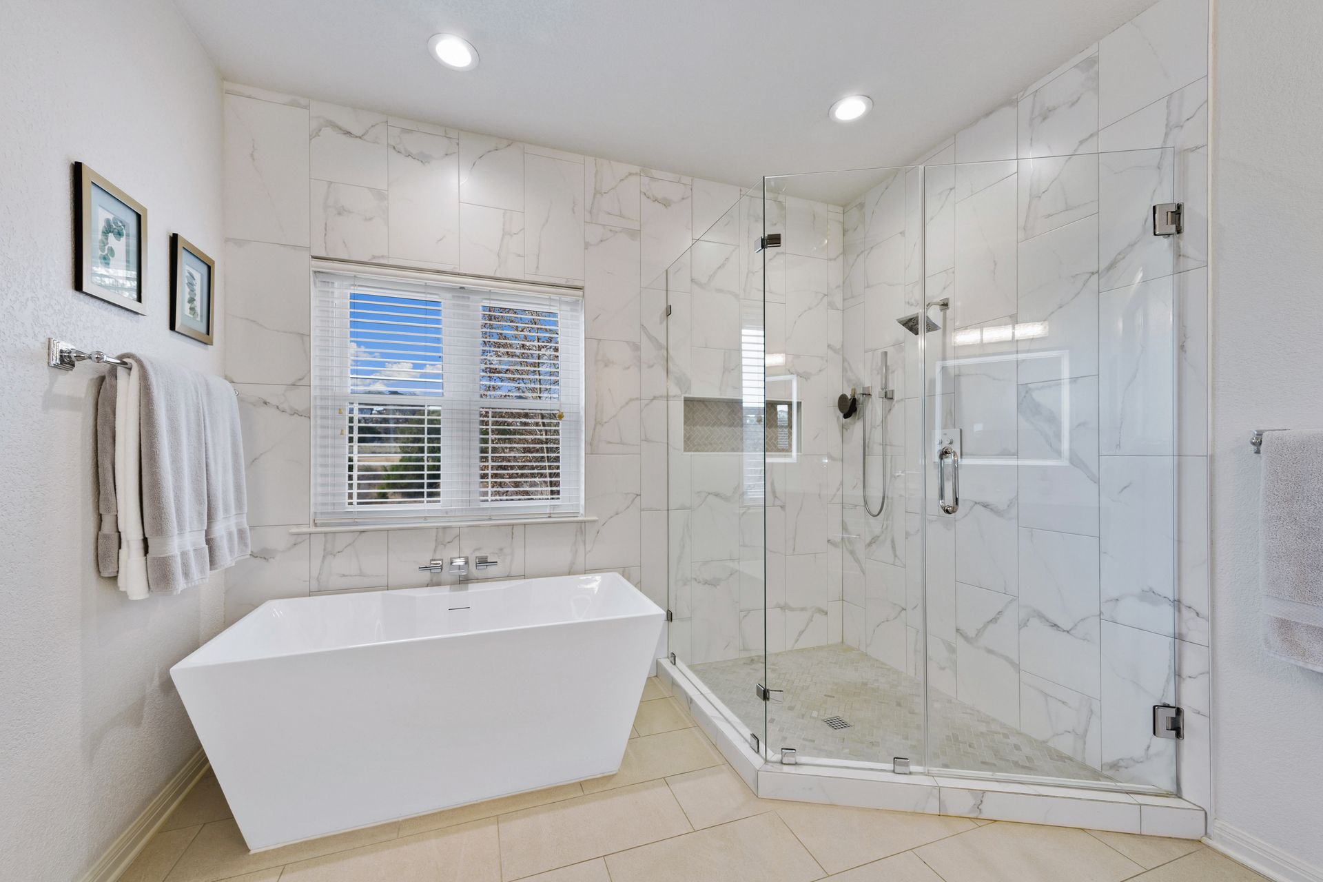 Bright modern bathroom with freestanding tub, glass shower, marble walls, and window blinds