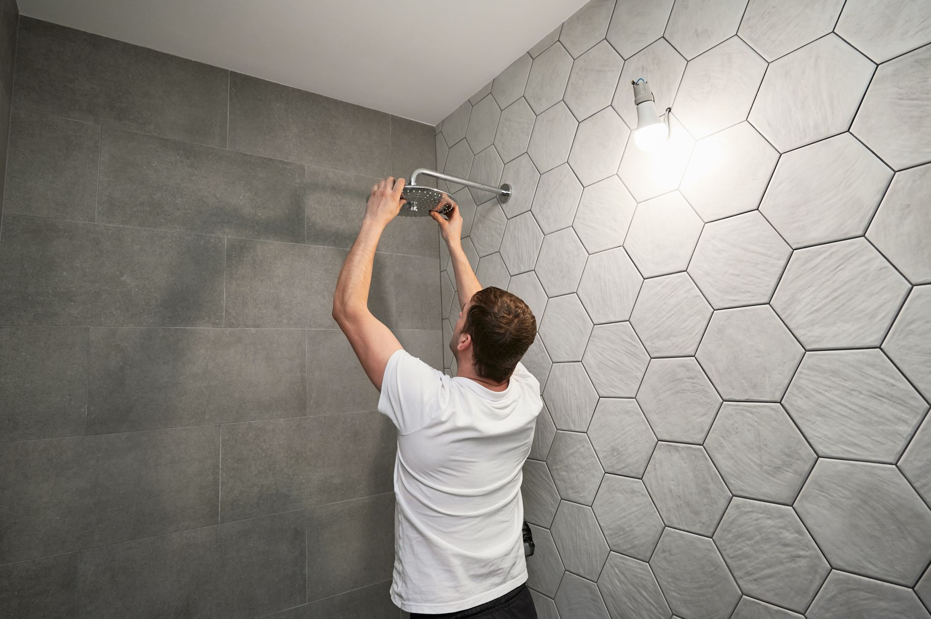 A contractor installing a shower head after putting grey hexagon tiles on a bathroom wall.