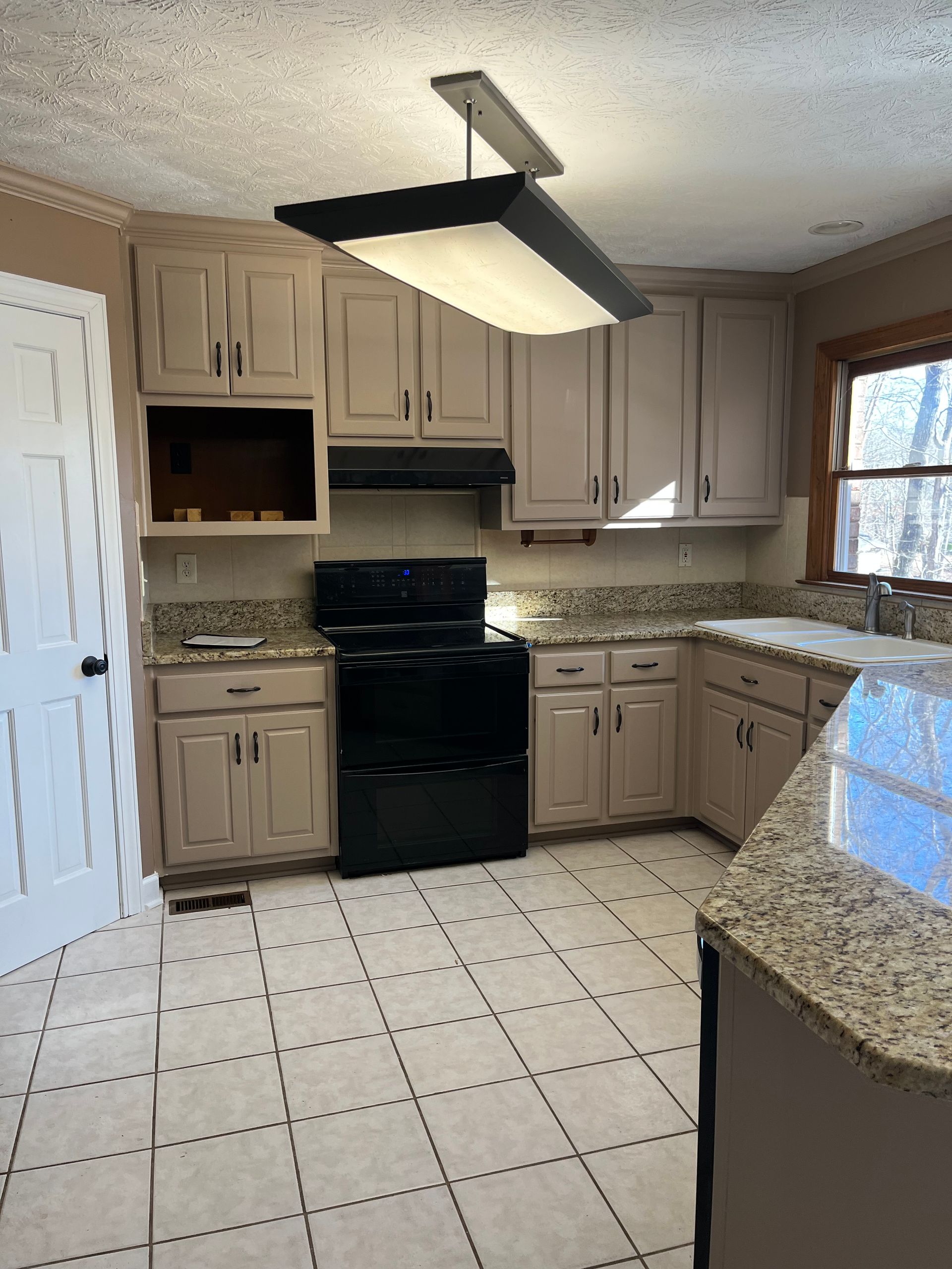 Compact kitchen with beige cabinets, black stove, tiled floor, and a ceiling light fixture.