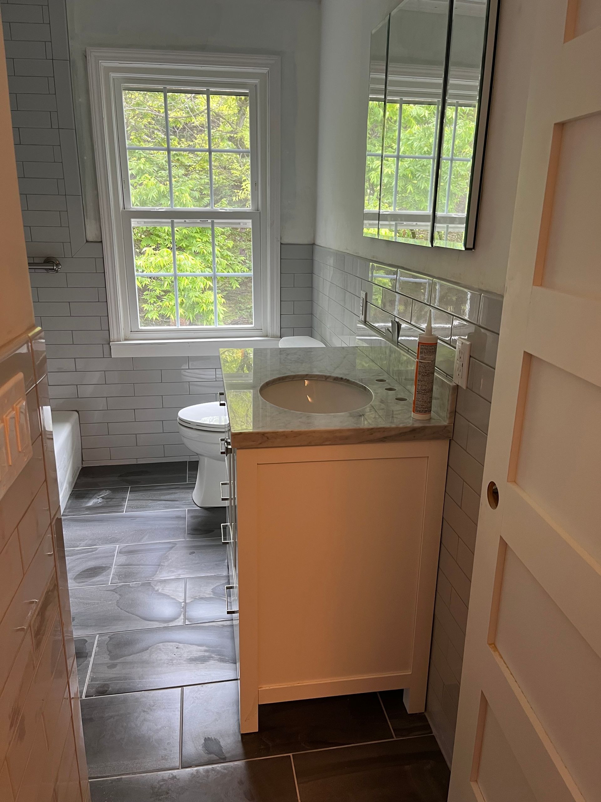 Small bathroom with white vanity, round sink, mirror, tiled floor, and two windows with greenery outside