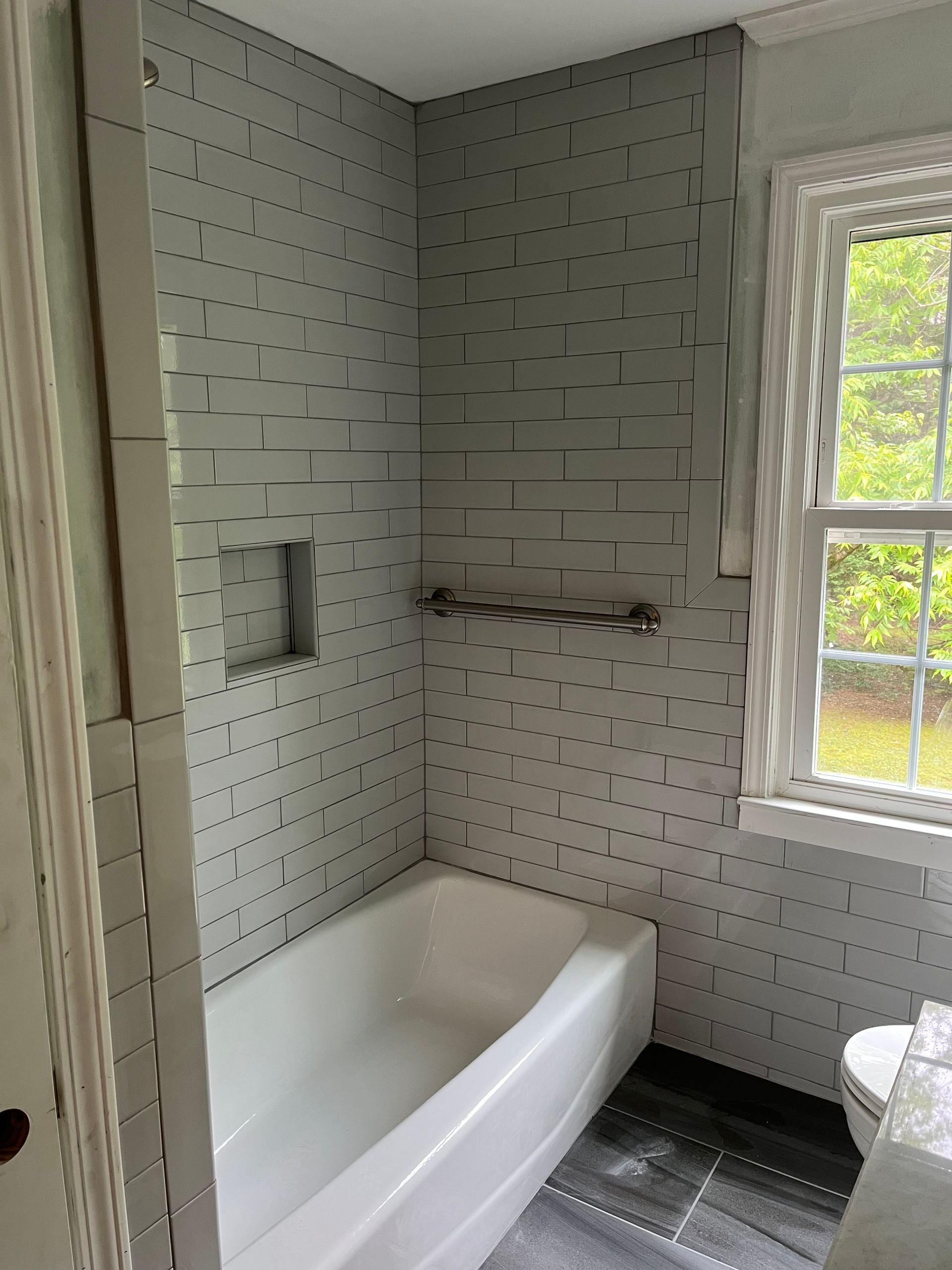 Small white-tiled bathroom with a white tub, black floor, and a window letting in daylight.