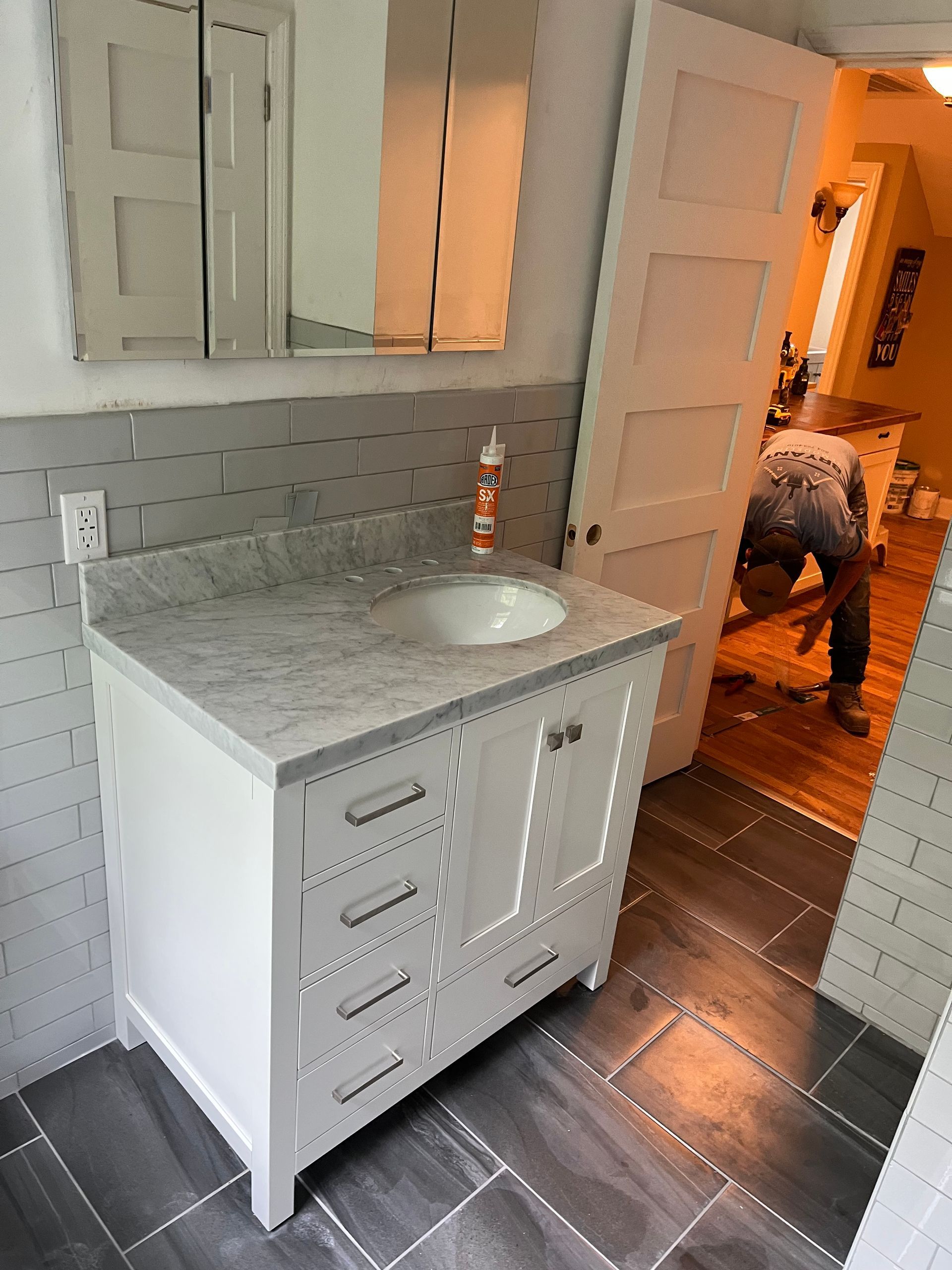 Small white bathroom vanity with mirror over dark tile floor; person visible in doorway in the background.