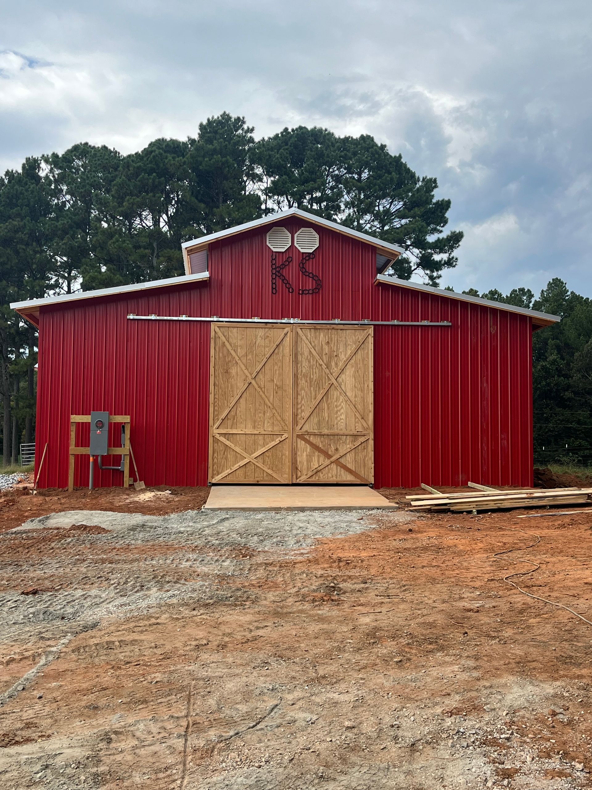 Red barn with large wooden double doors on a gravel lot under a cloudy sky.