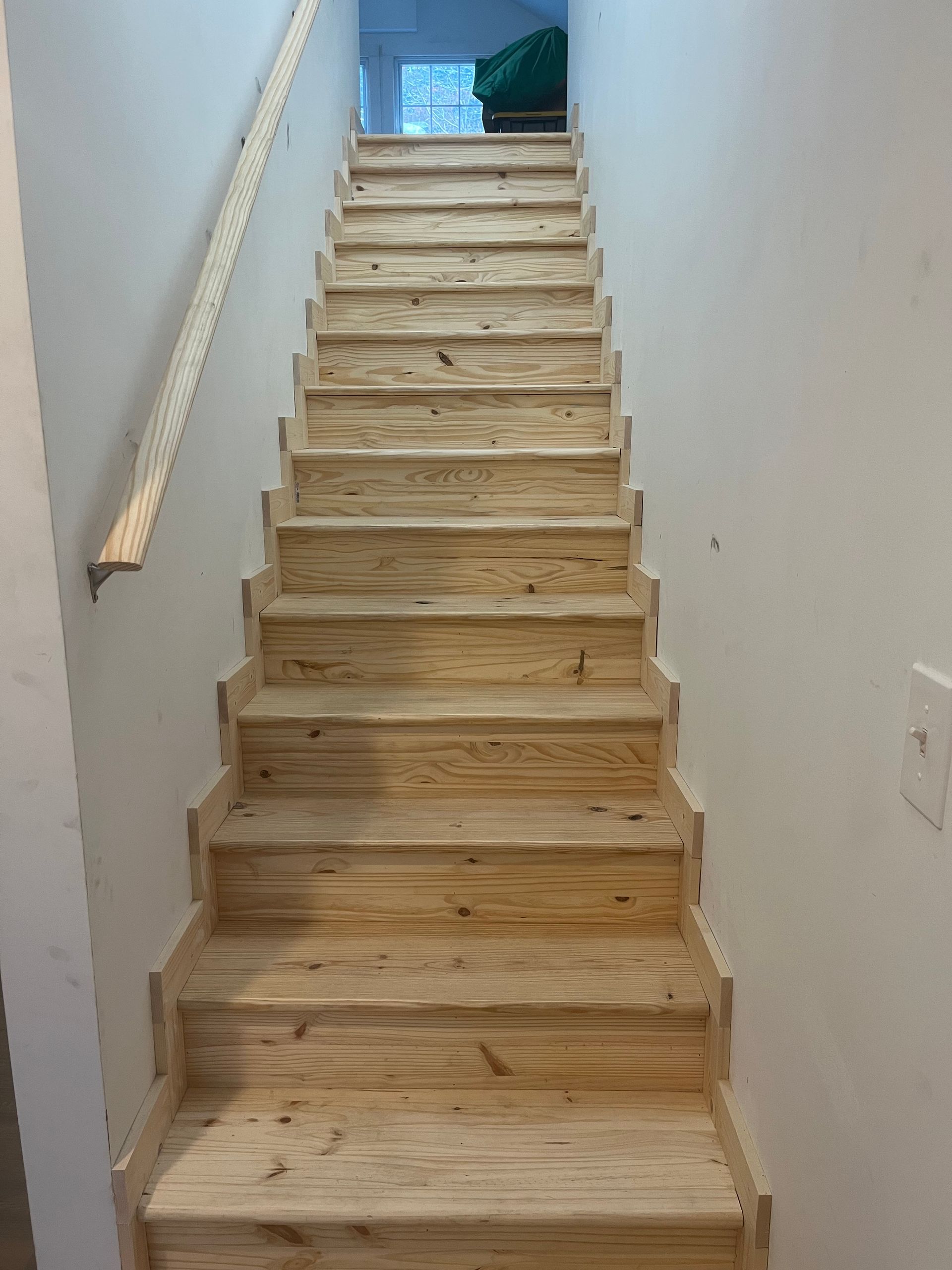 Narrow unfinished wooden staircase with white walls and a handrail, leading up to a bright landing.