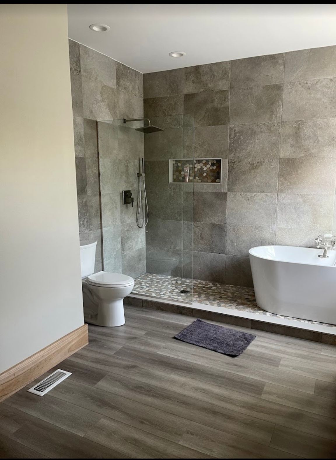 Modern bathroom with gray tiled walk-in shower, white toilet, and freestanding tub on wood-look floor
