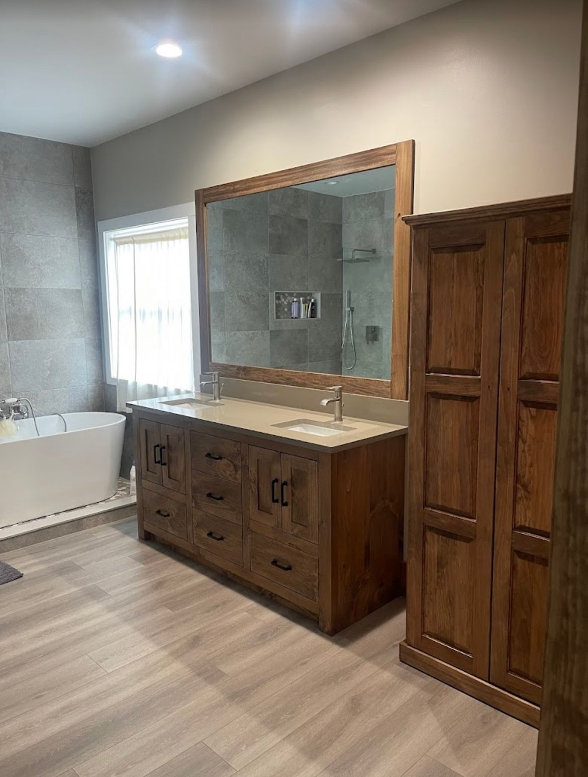 Modern bathroom with double vanity, large mirror, bathtub, and wooden storage cabinet on the right.