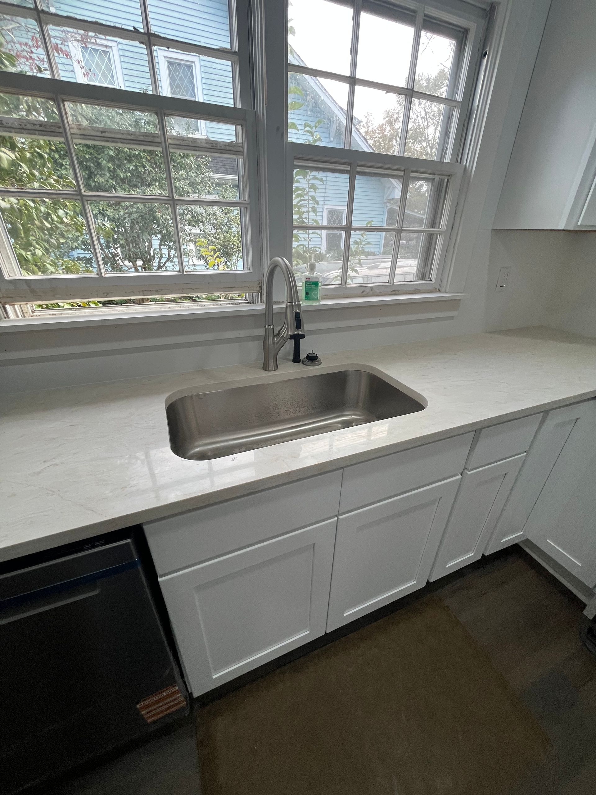 White kitchen sink beneath large windows, with white countertop and cabinets.