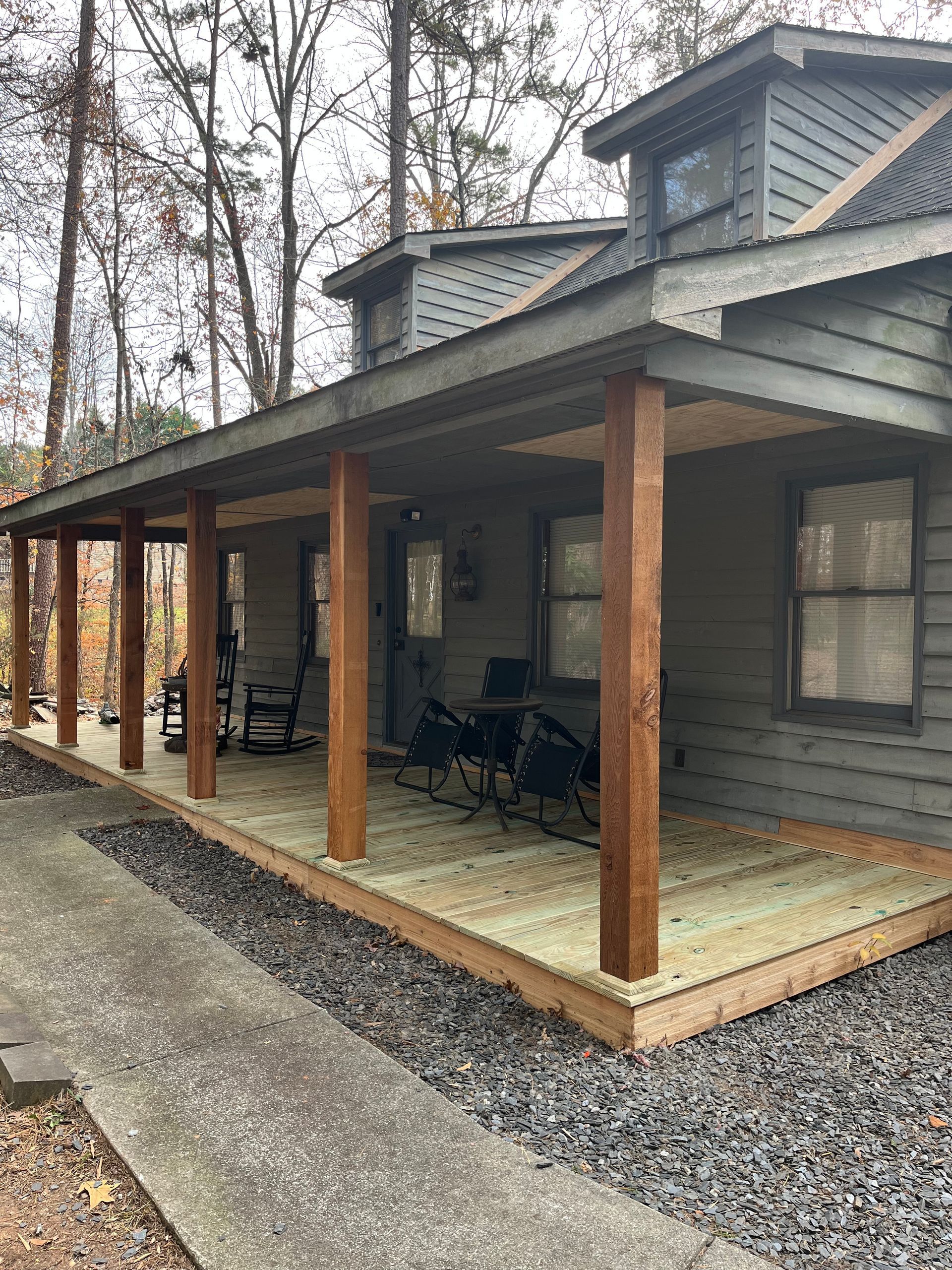 Covered porch with wooden posts and chairs on a gray house, beside a gravel driveway and wooded yard.