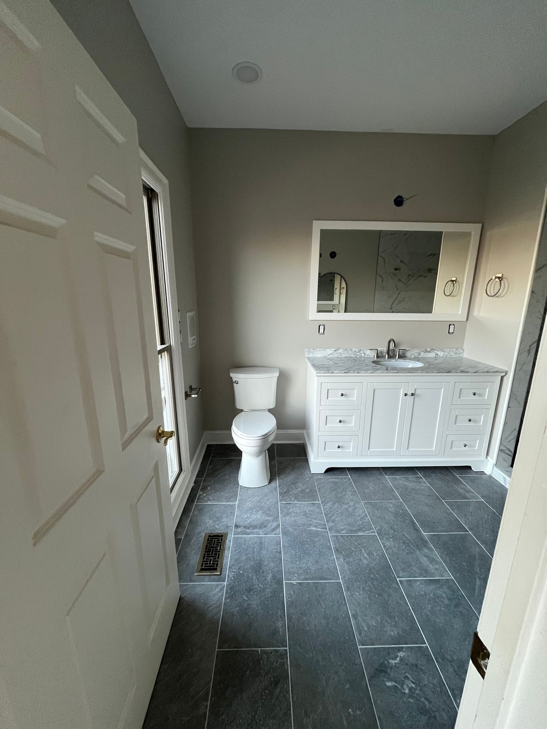 Empty beige bathroom with white vanity, mirror, toilet, and dark tile floor.