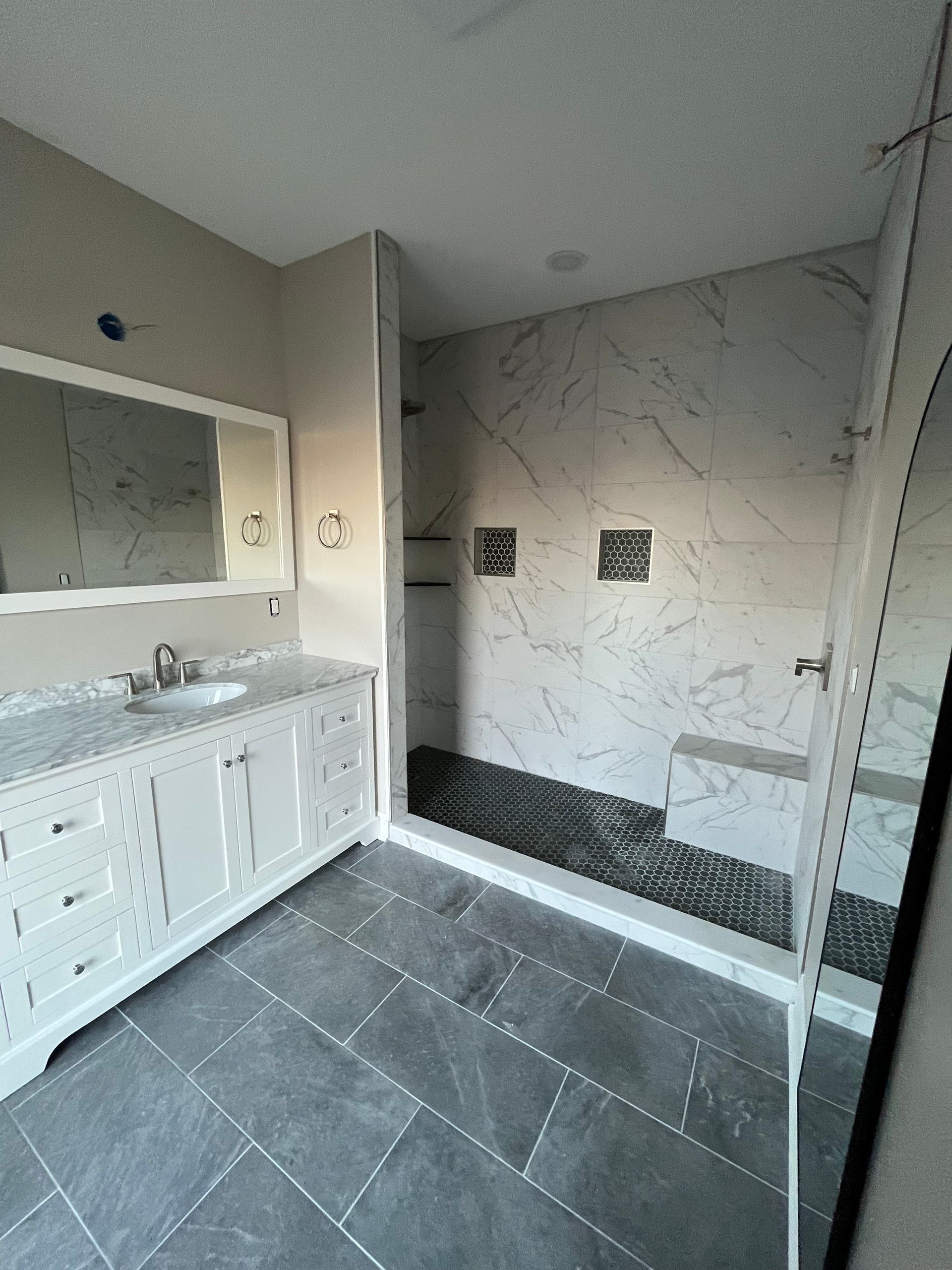 Modern bathroom with white vanity, large mirror, dark floor tiles, and a marble-look shower area.