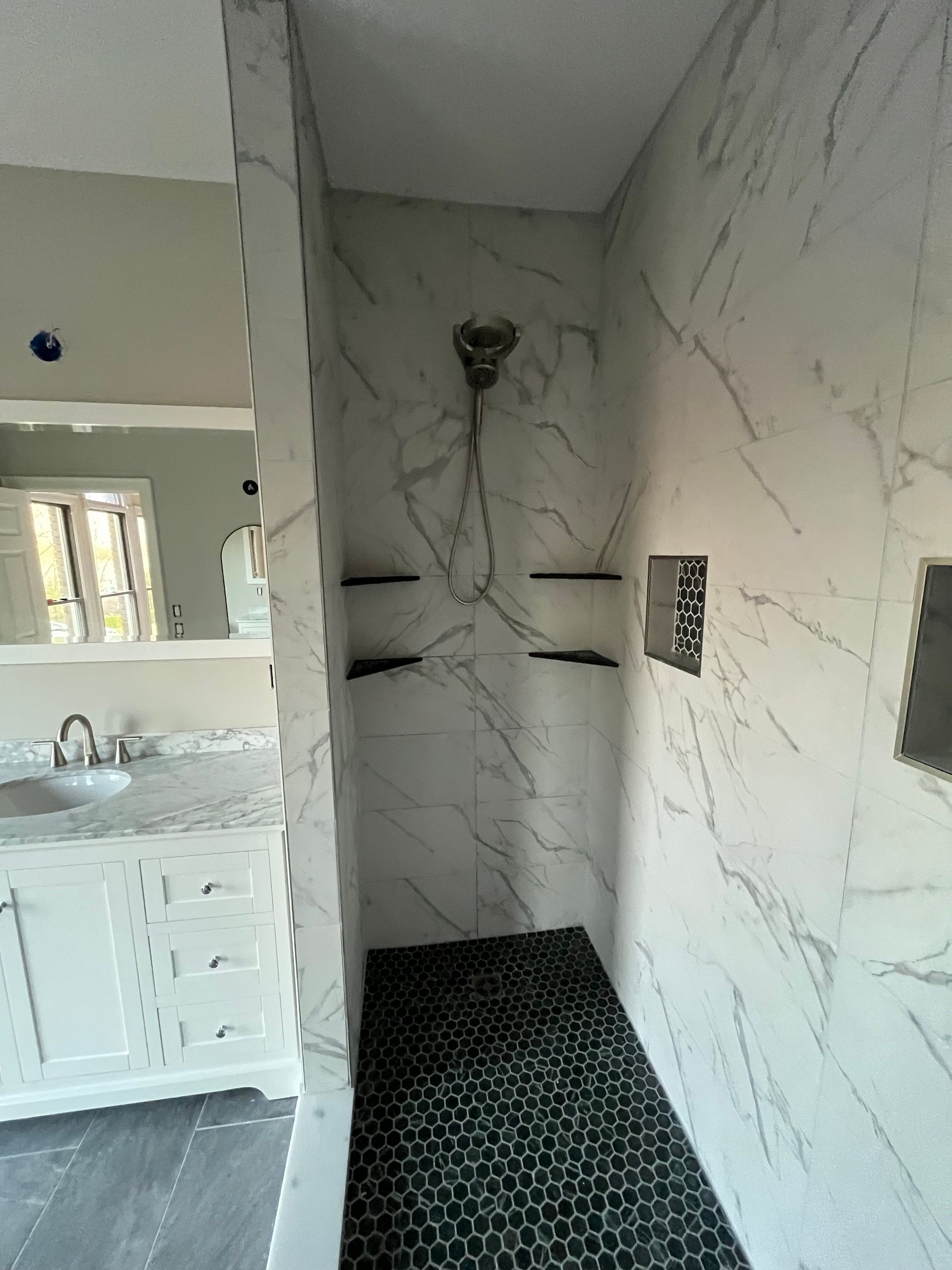 Tiled walk-in shower with black floor tile, marble-look walls, and white vanity visible nearby