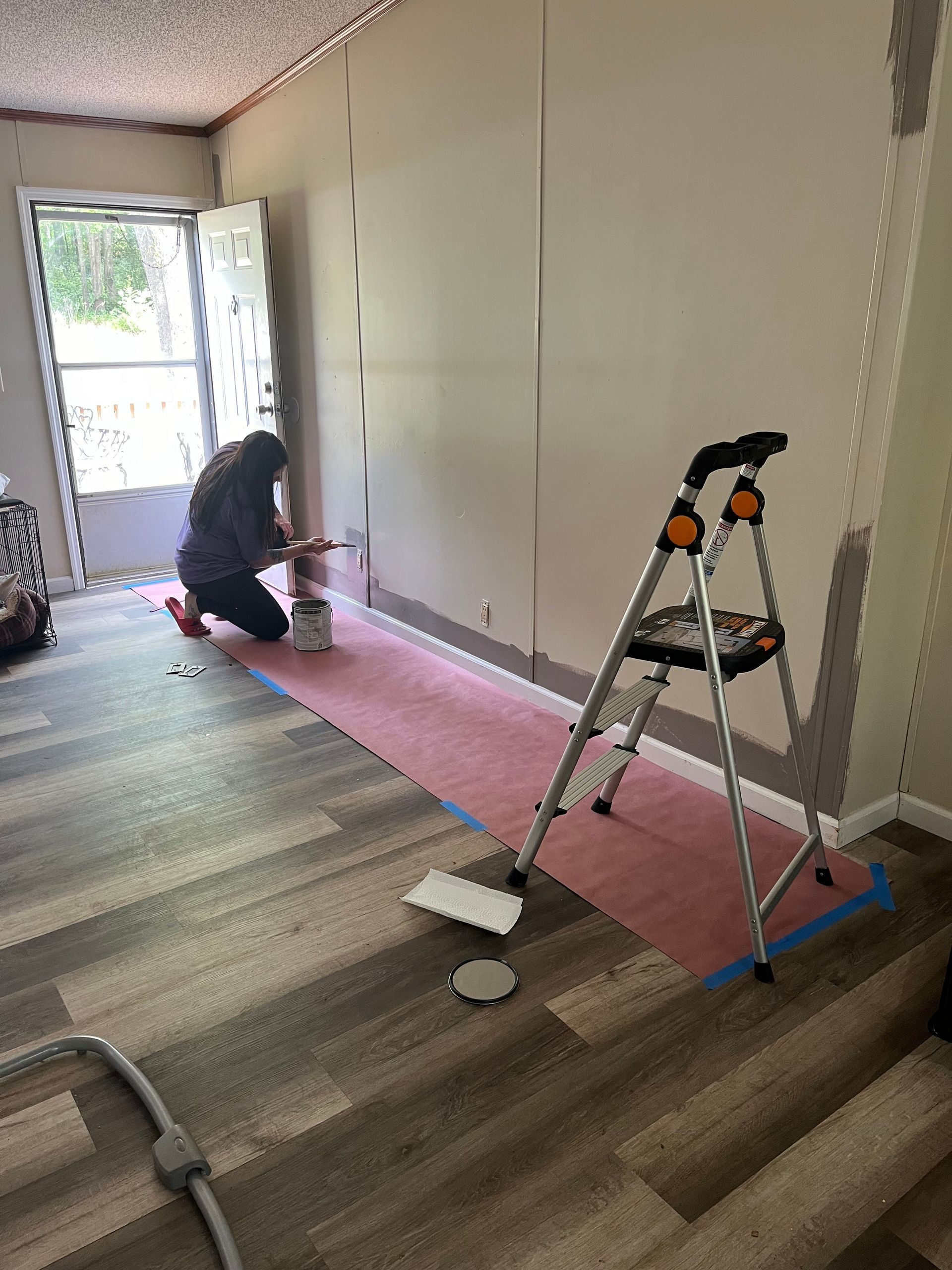 Person painting a wall pink while standing on a step ladder in a room with wood flooring.