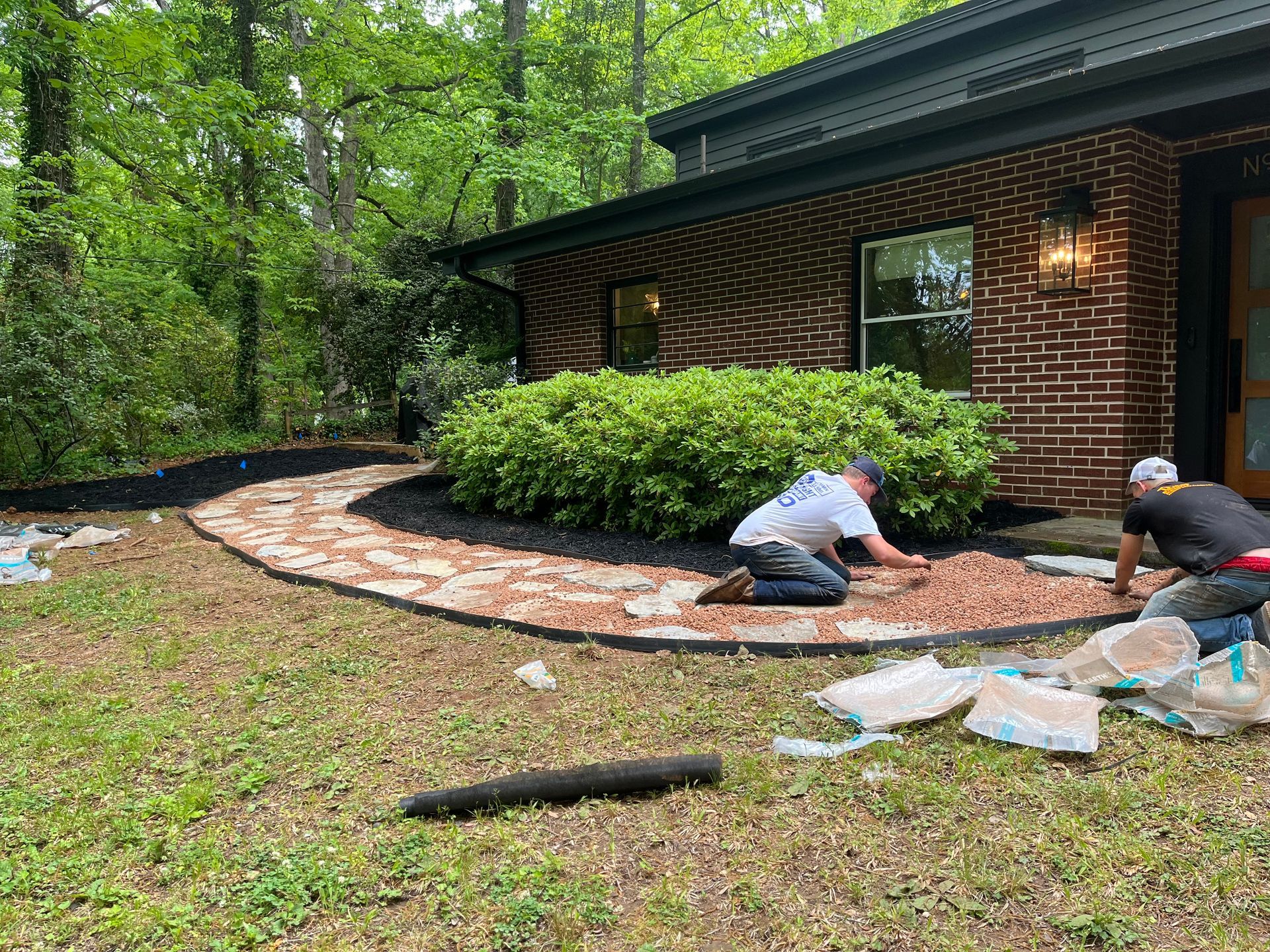 Two workers installing edging around a curved flower bed beside a brick house in a wooded yard