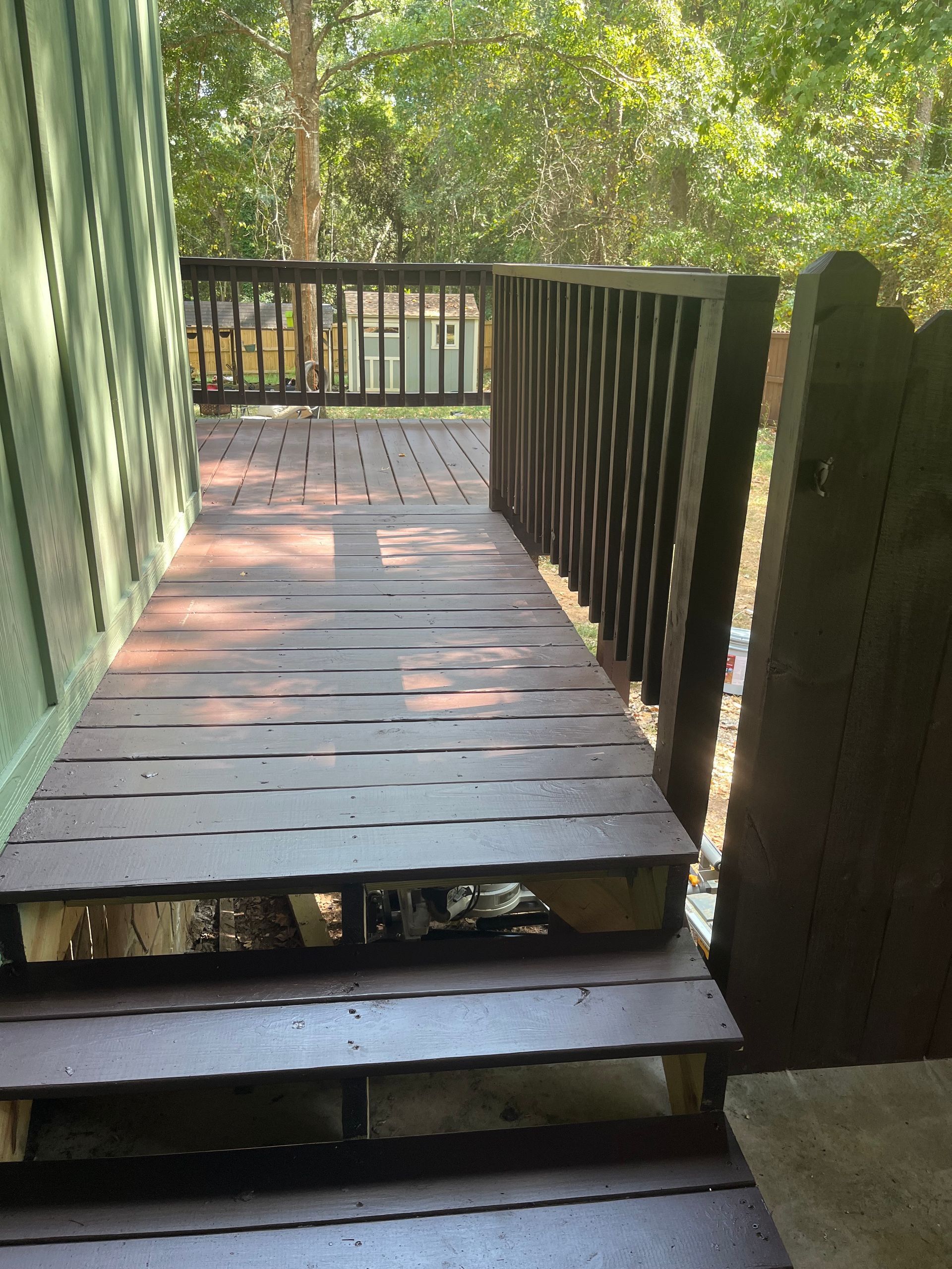 Weathered wooden porch walkway with railings leading to a deck beside a green house.