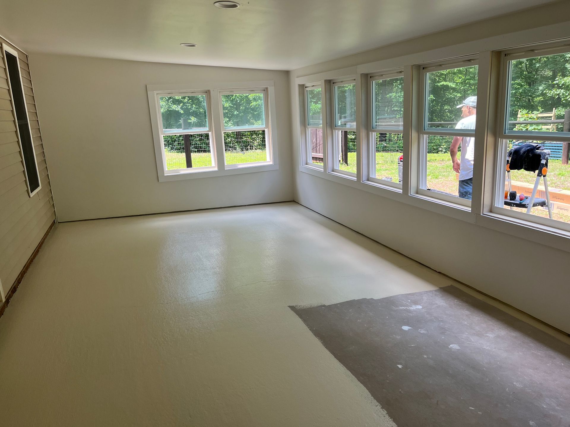 Empty sunlit room with white walls, large windows, and a beige floor, with a gray patch near the corner