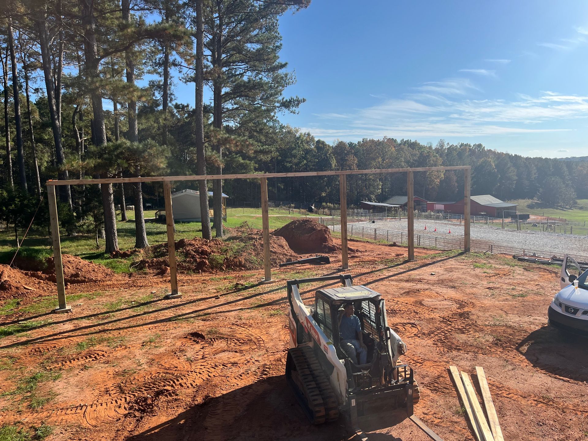 Cleared dirt lot with a wooden frame, trees, gravel road, and a skid steer in the foreground