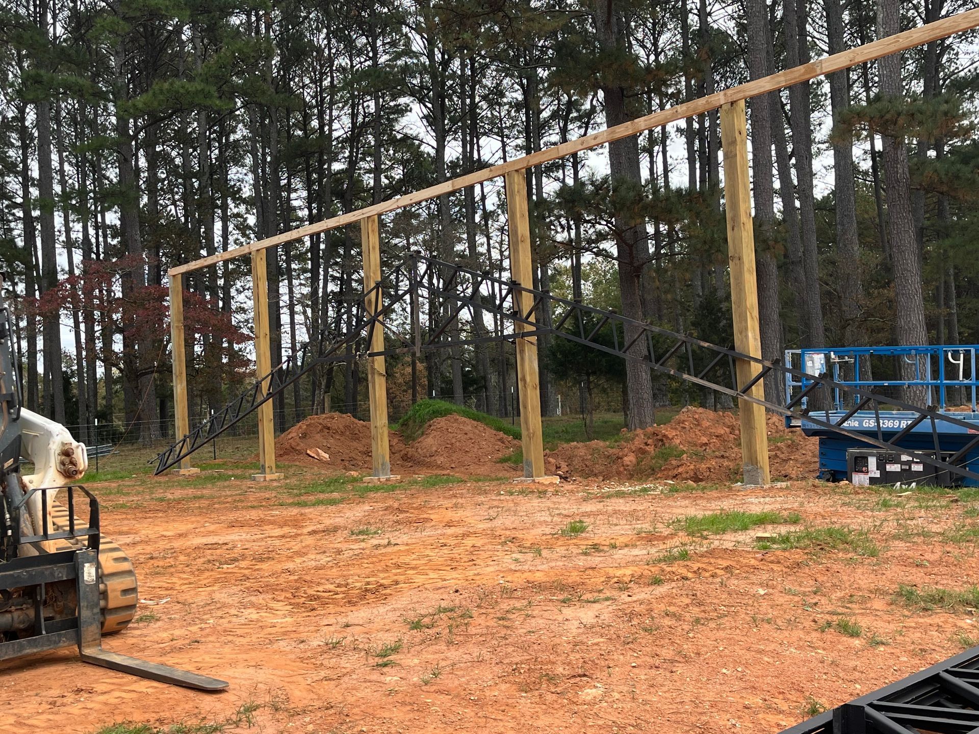 Construction site with wooden framework posts in a wooded area, muddy ground, and machinery on the left and right.