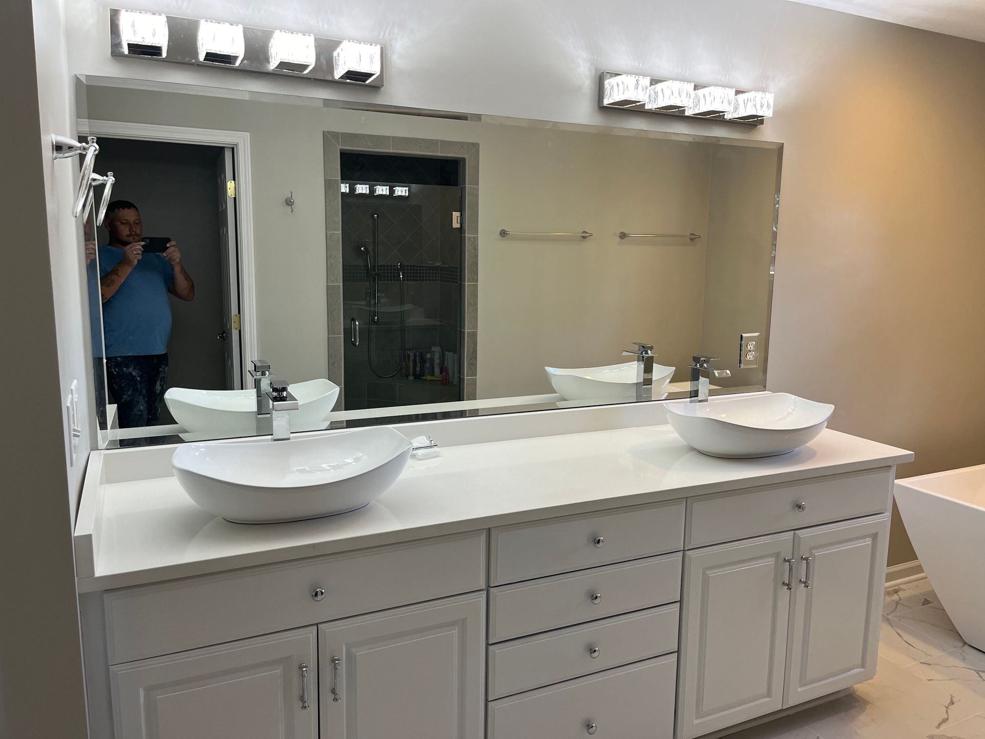 Bathroom vanity with large mirror, dual sinks, and wall lights; a person is reflected taking the photo.