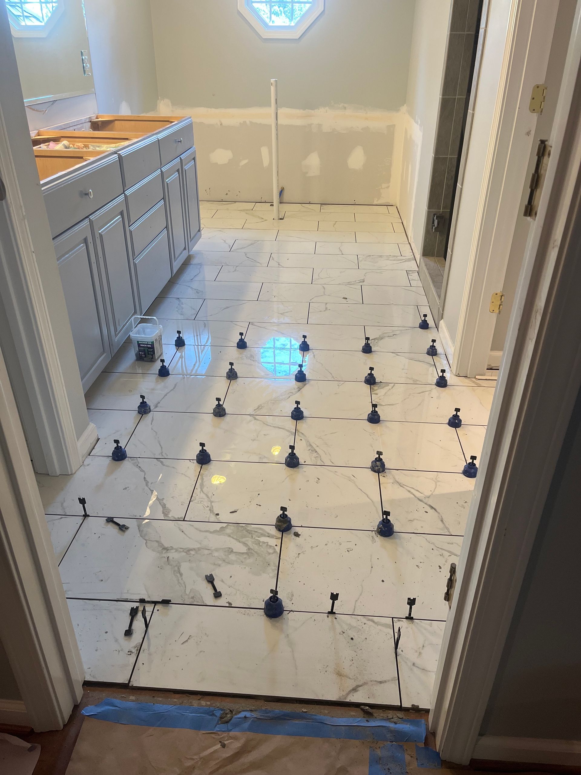 Bathroom floor under renovation with white tiles, blue spacers, and cabinet vanity along the left wall