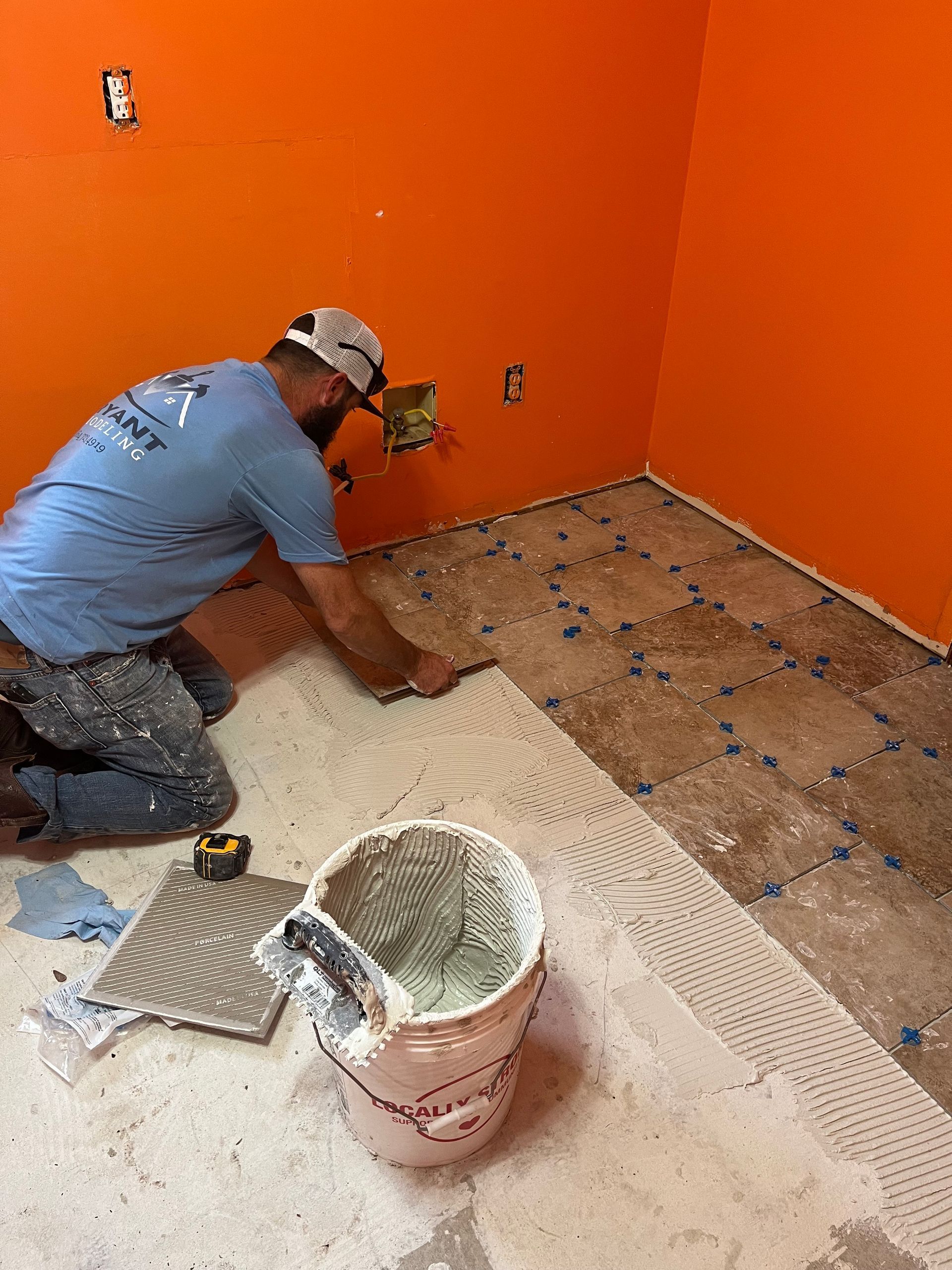 Worker installing tile in orange-walled room, with bucket and trowel on the floor.