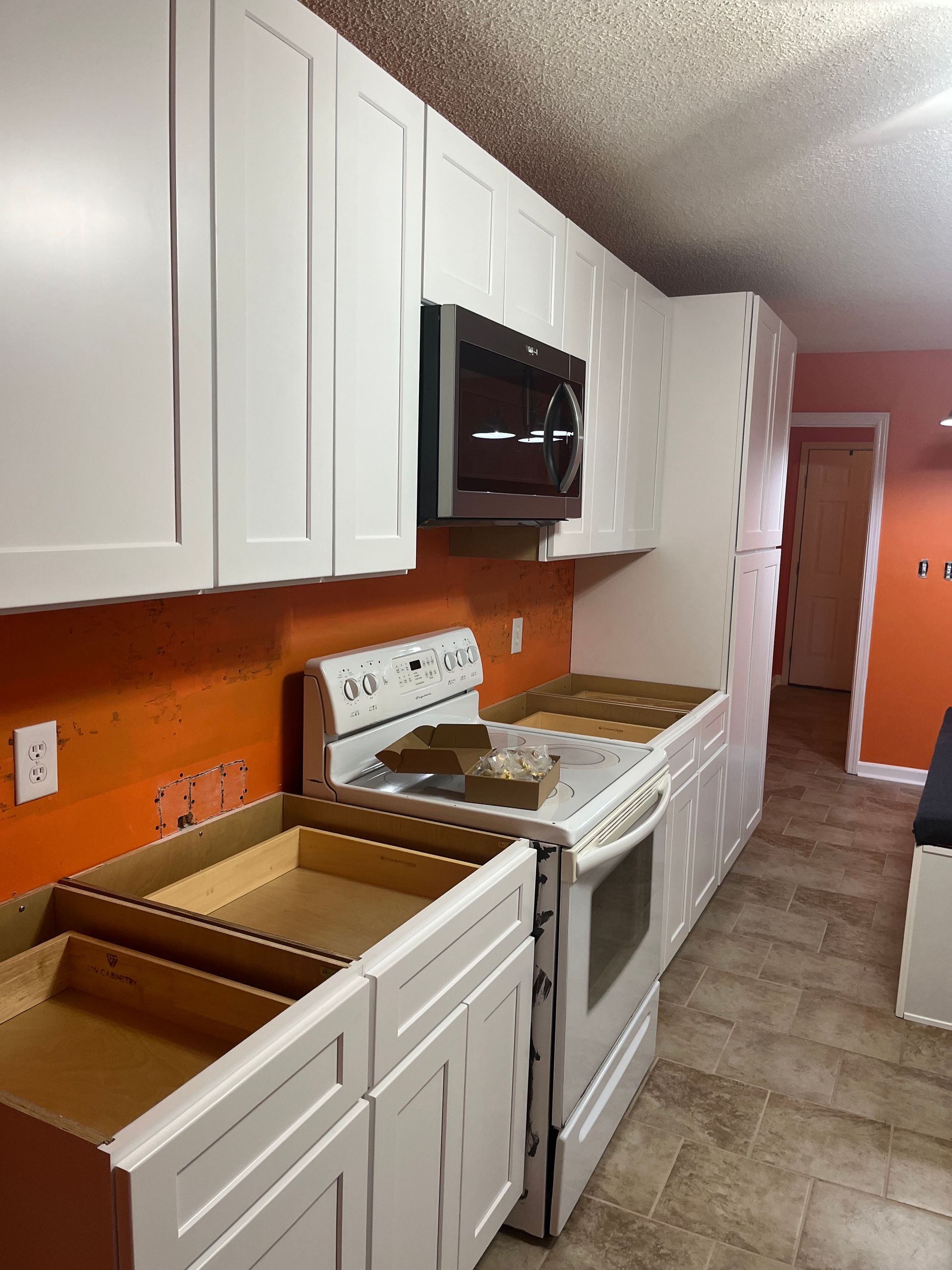 White kitchen with orange walls, white cabinets, stove, microwave, and open drawers along a narrow hallway.