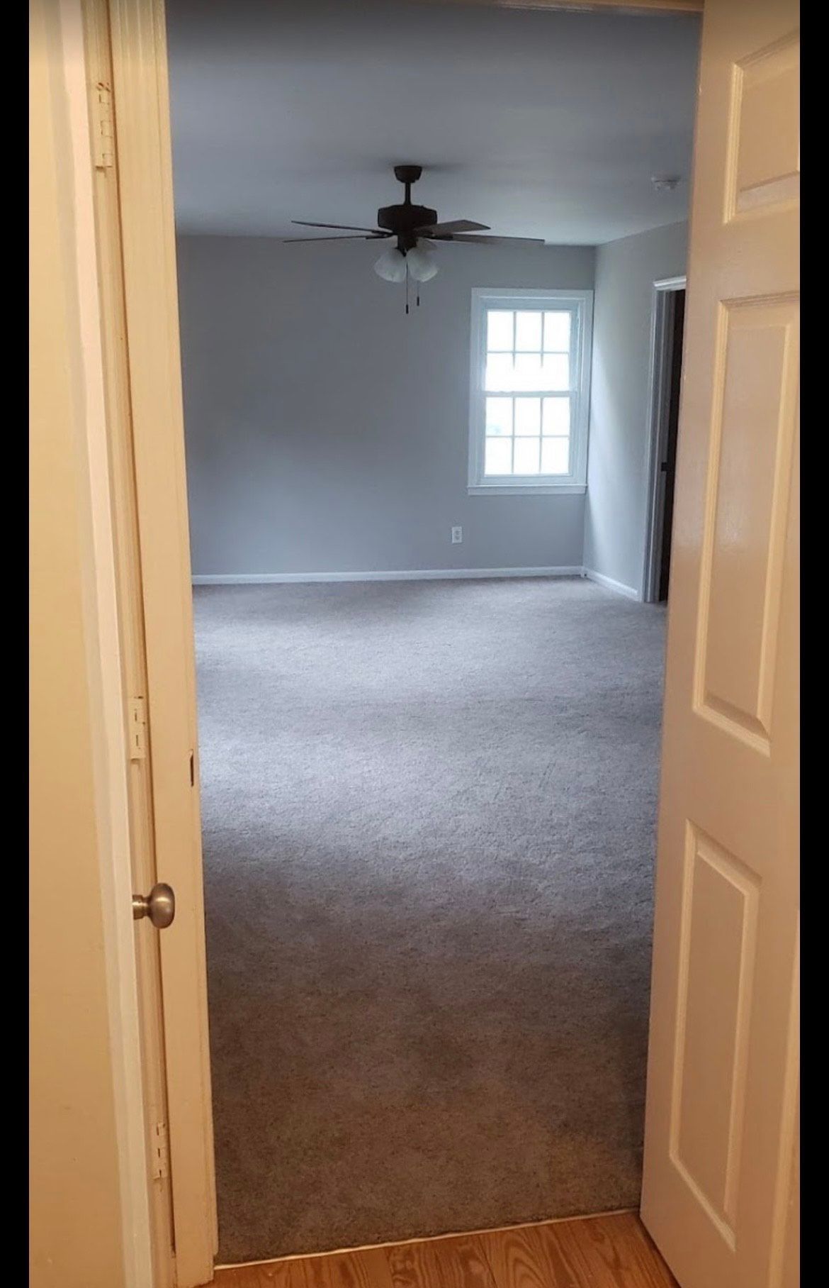 Open doorway to an empty gray-carpeted room with a ceiling fan and window.