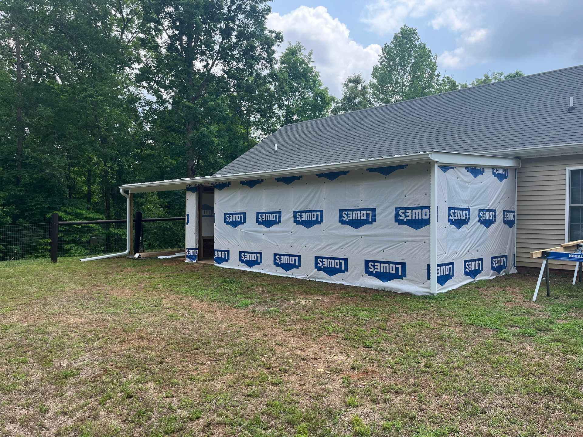 House exterior under construction with housewrap on the porch, beside a wooded yard