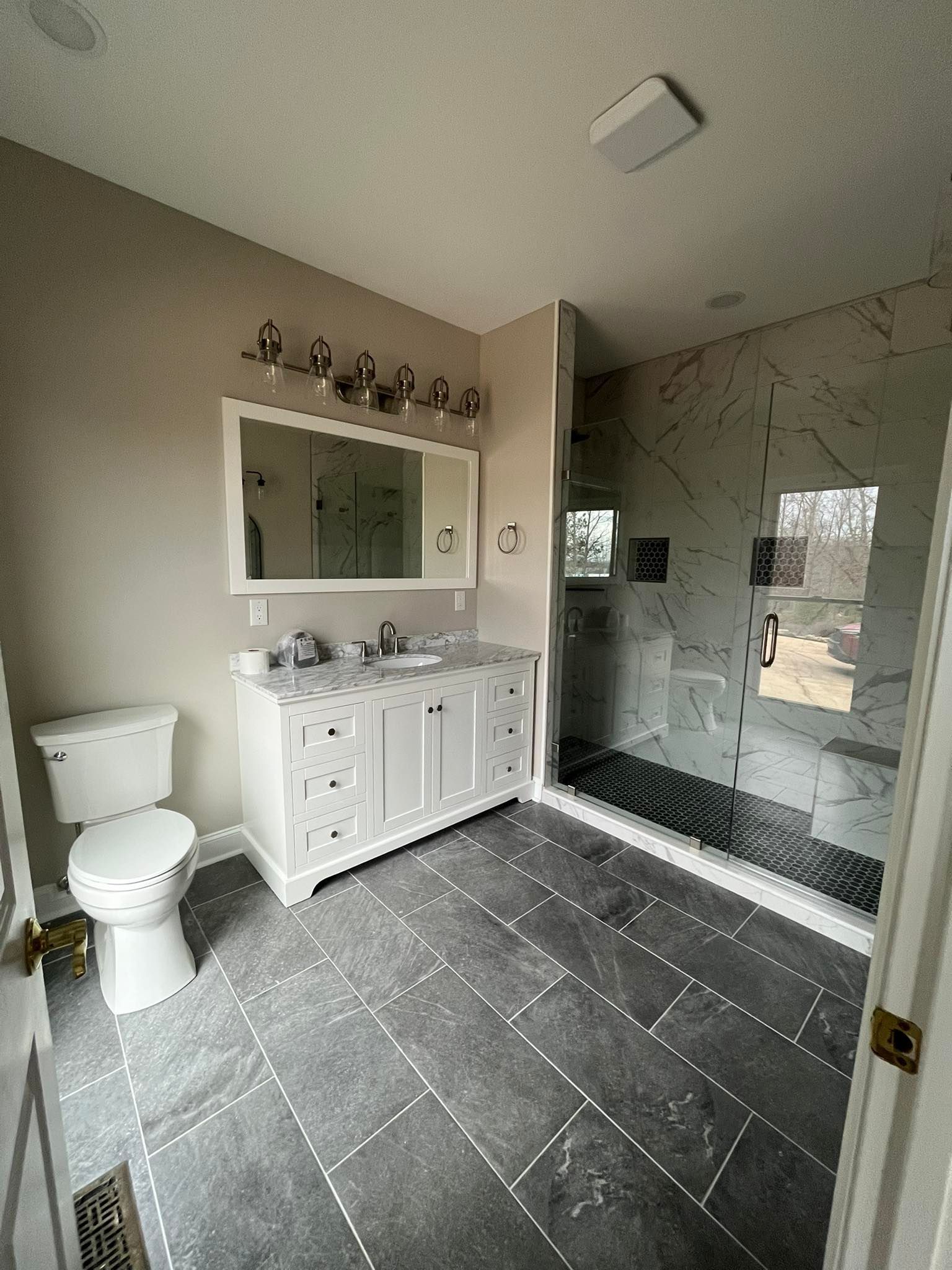 Small modern bathroom with toilet, vanity sink, mirror, and glass shower enclosure on gray tile floor.