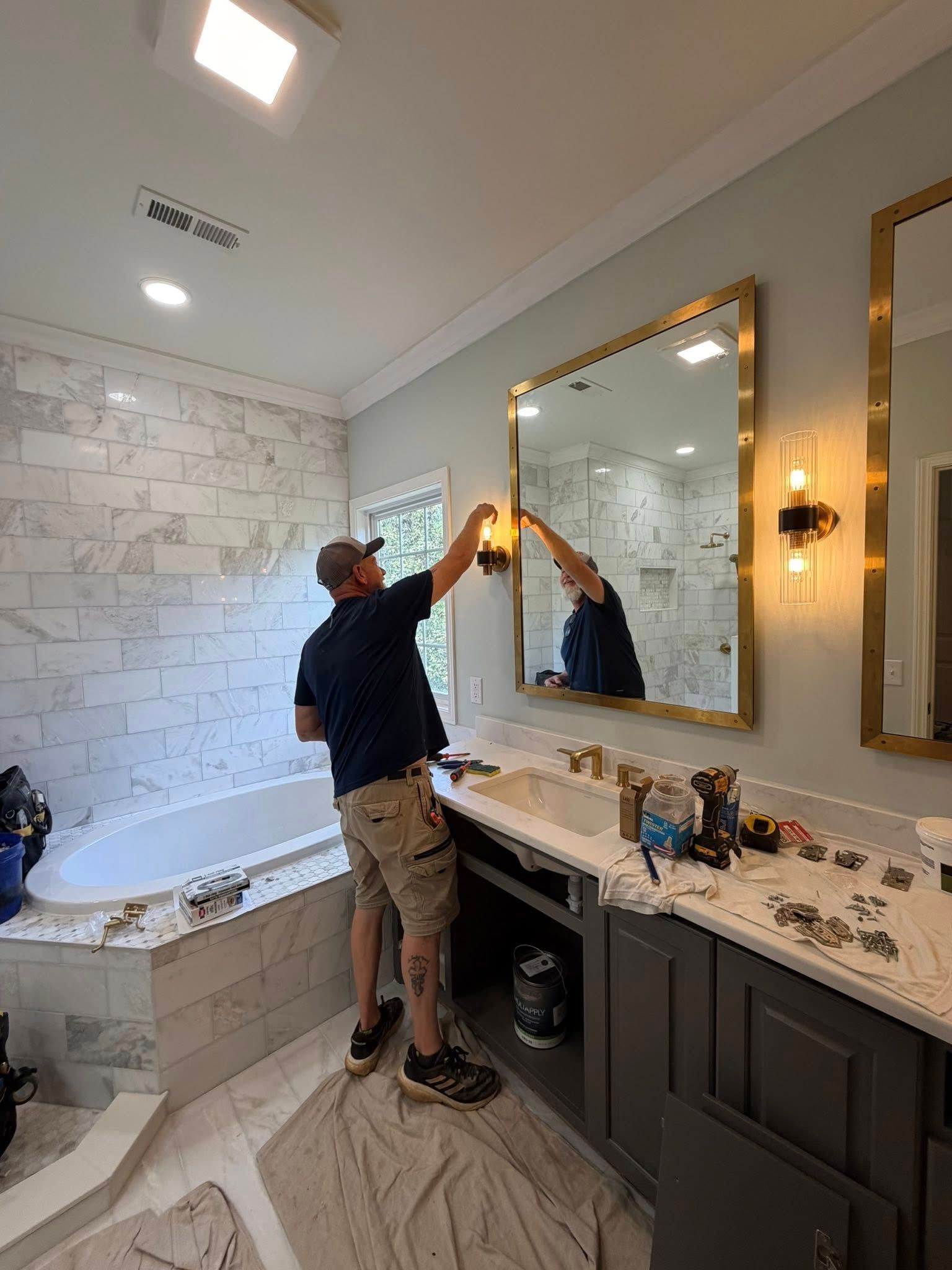 Worker installing a vanity light in a bathroom with gold-framed mirrors and white tile.