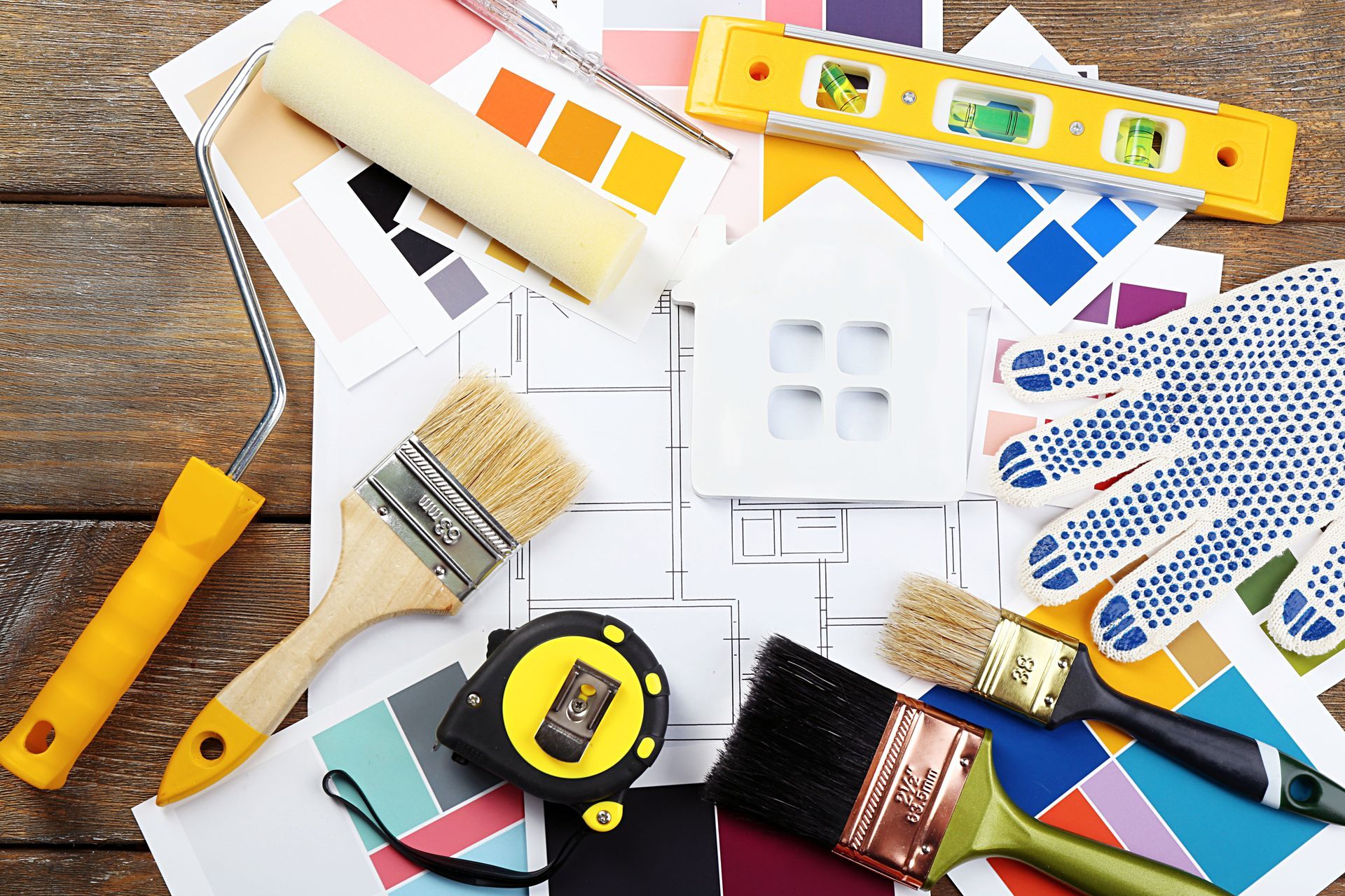 A bunch of paint brushes , gloves , a tape measure , and a house on a wooden table.
