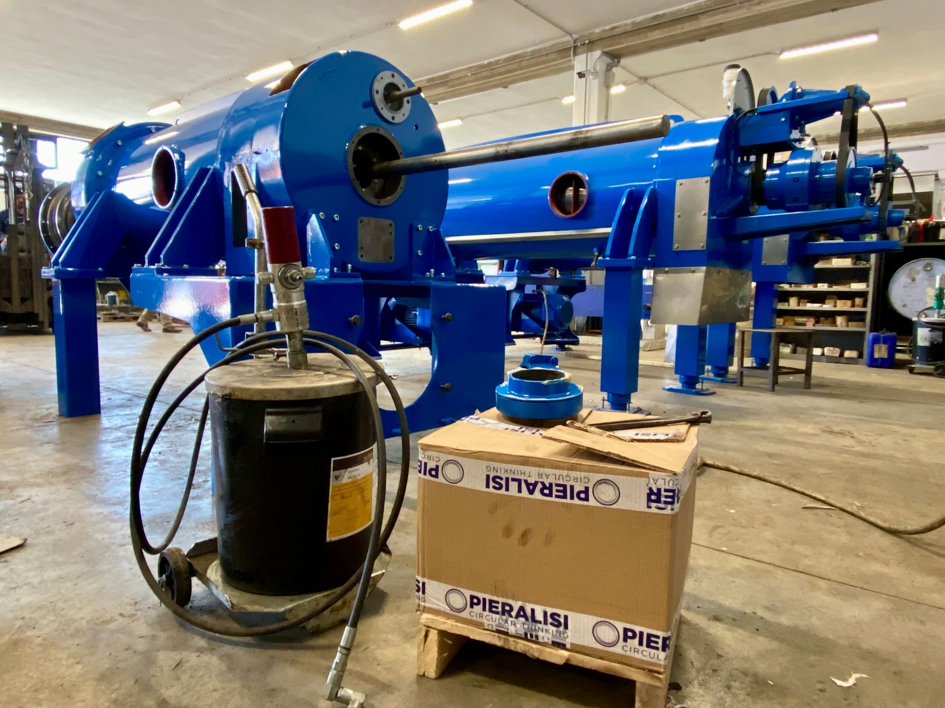 Macchinari industriali blu in un'officina, con tubi flessibili e un serbatoio nero in primo piano.