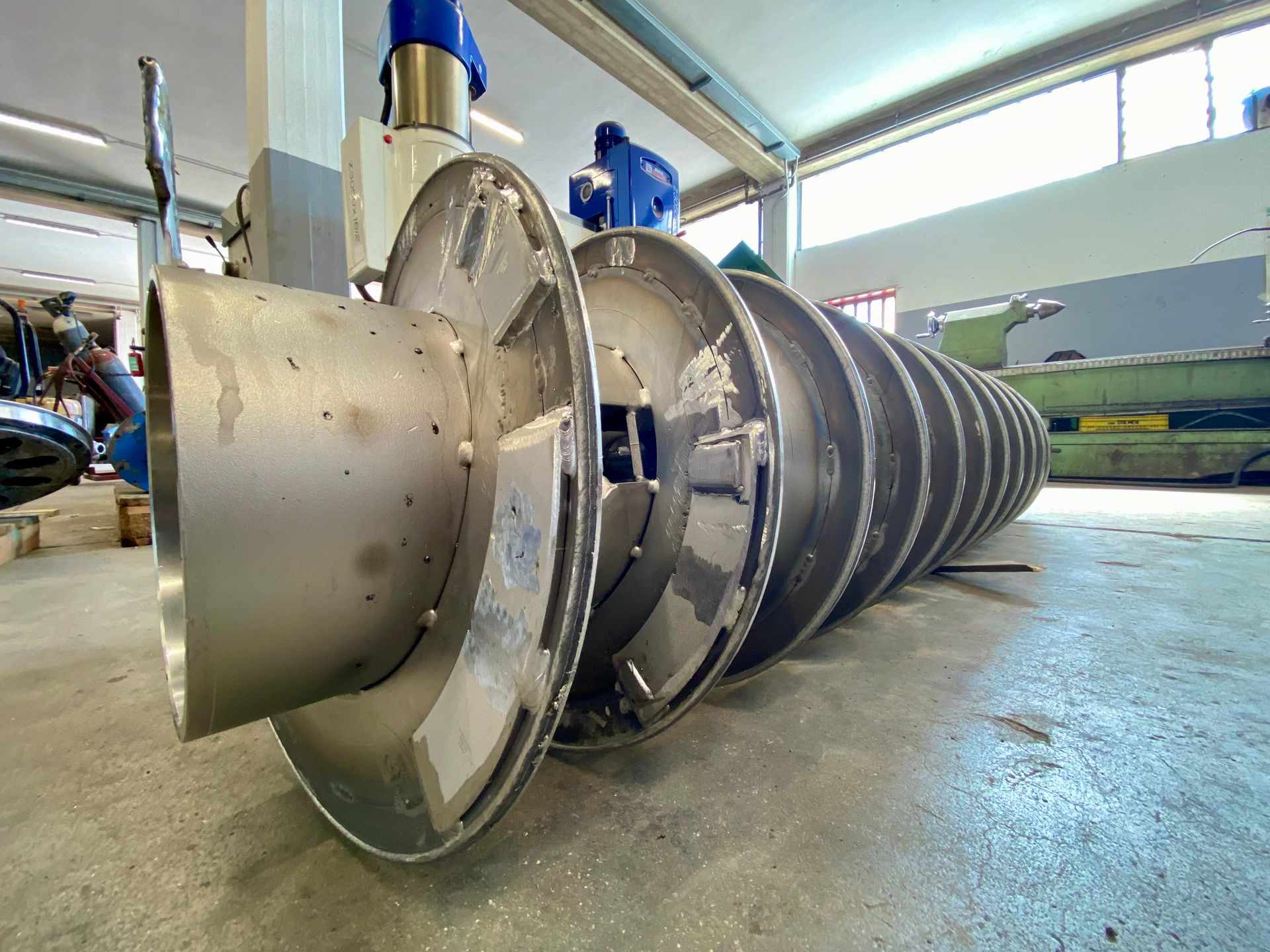 Un grande rotore di turbina in metallo giace sul pavimento di un'officina industriale.