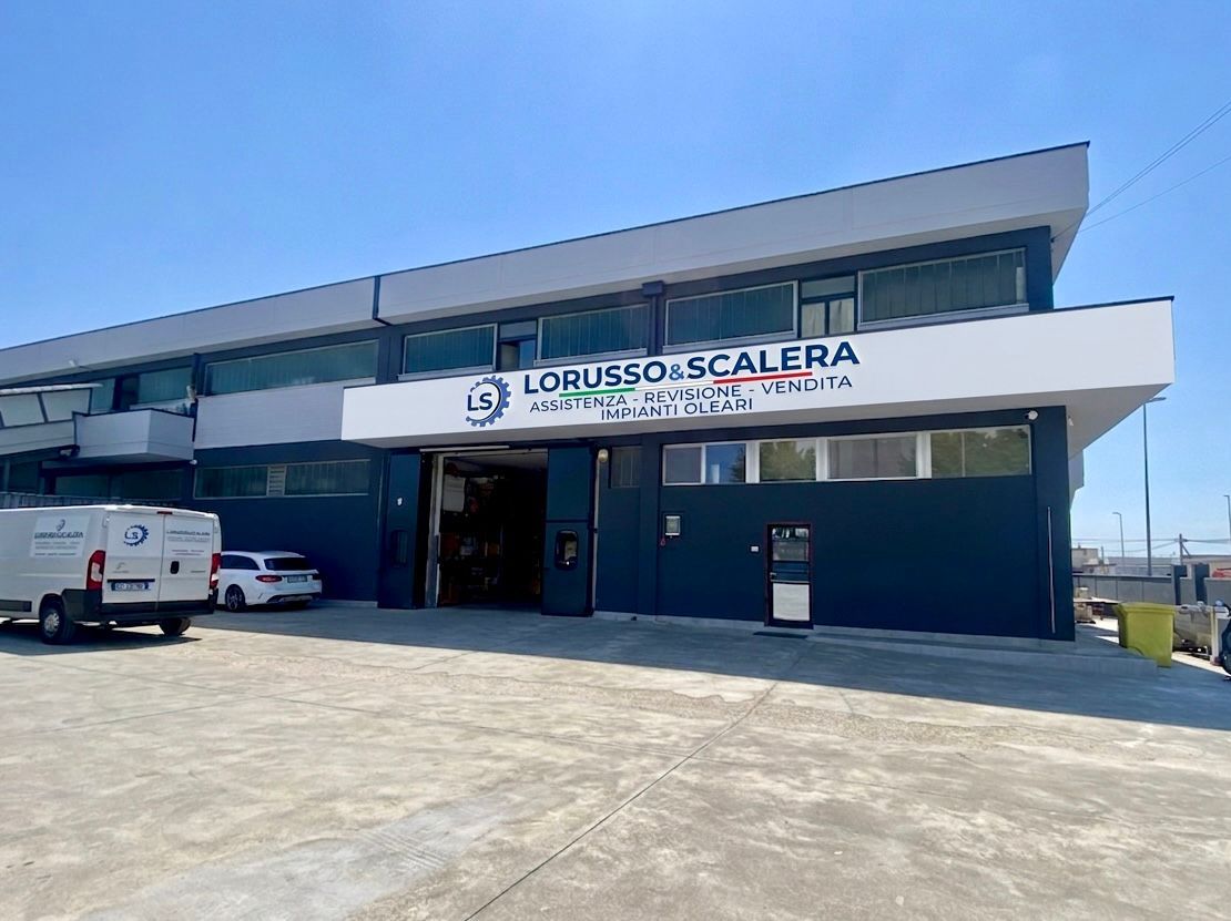 Esterno di un edificio commerciale blu e bianco con un camion bianco parcheggiato e cielo soleggiato.