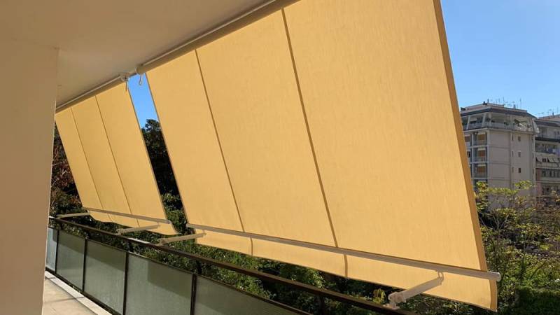 Tende da sole retrattili gialle su un balcone, che ombreggiano la terrazza di un edificio con vista sulla città.