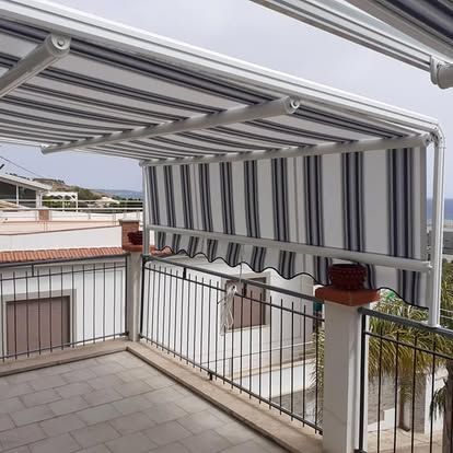 Balcone coperto con tenda da sole retrattile a righe, ringhiera e pavimento piastrellato, con vista sugli edifici e sugli alberi circostanti.