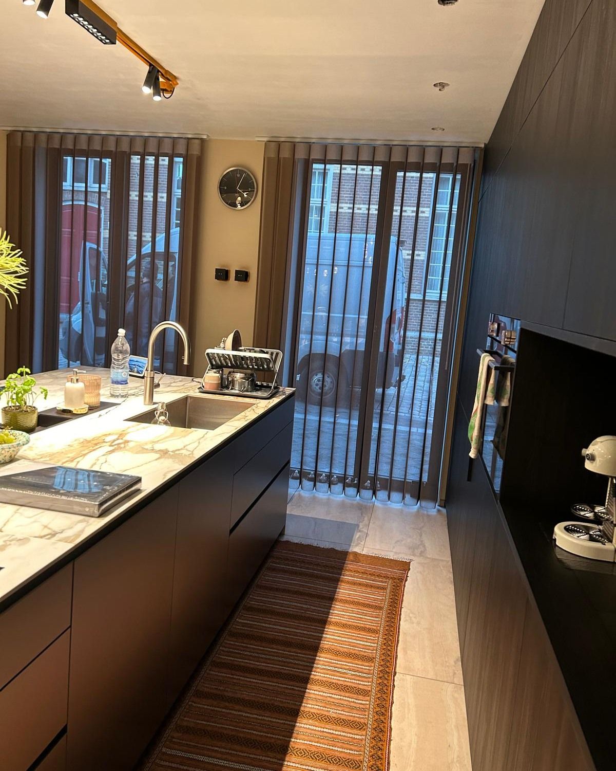 Cucina moderna con mobili scuri, lavello a isola, pavimento in legno massello e porte scorrevoli in vetro con tende trasparenti.