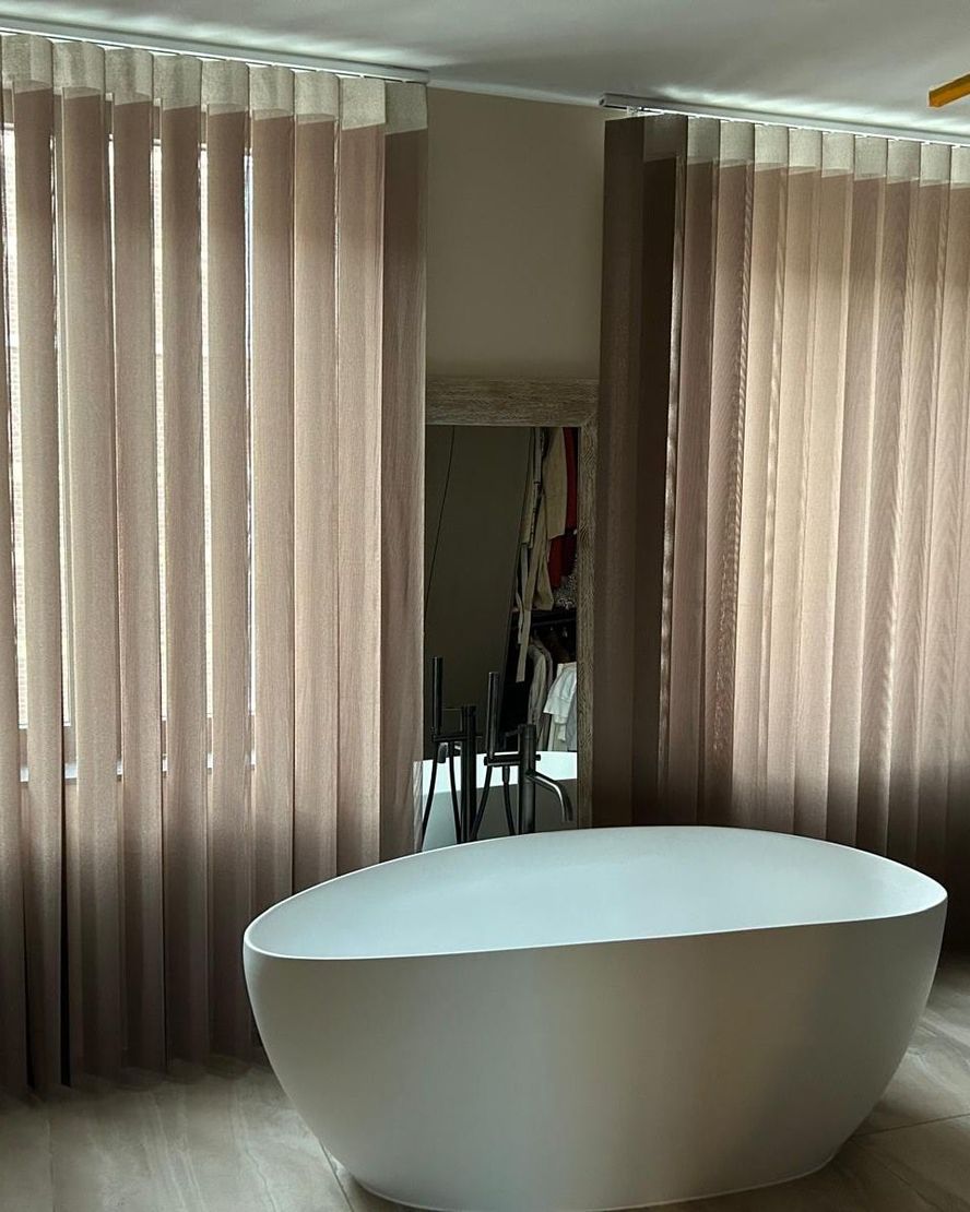 Camera moderna con tende beige e vasca da bagno bianca freestanding posizionata al centro, vicino a uno specchio.