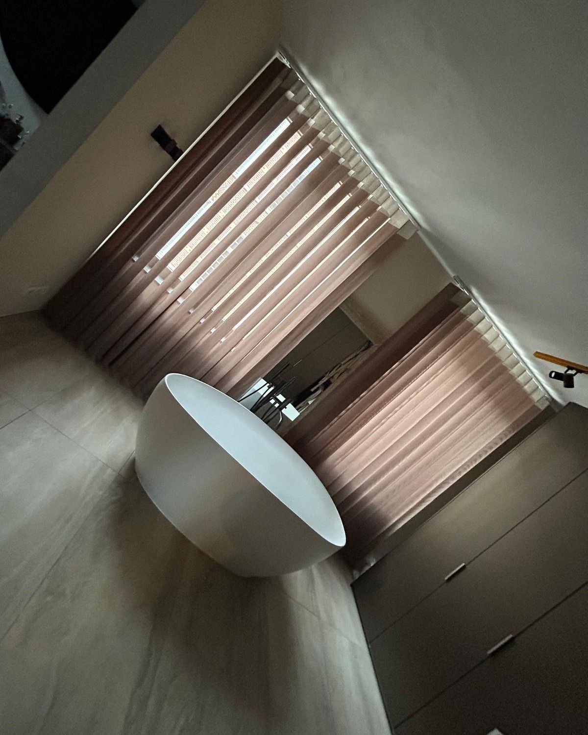 Stanza moderna con finestre alte, tende beige e un tavolo ovale bianco su un pavimento grigio.