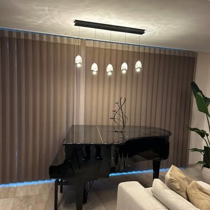 Un pianoforte a coda nero sotto lampade a sospensione in un soggiorno con tende beige e un divano bianco.