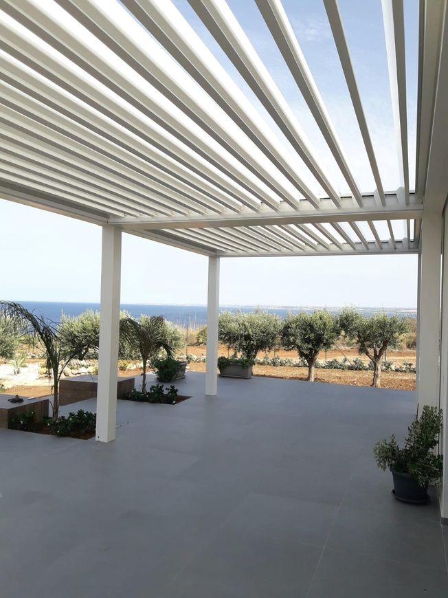 Patio moderno con pergola bianca, pavimento grigio, piante in vaso e vista mare oltre gli ulivi.