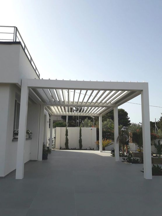 Patio moderno con pergola bianca, pavimento in cemento e vegetazione circostante sotto un cielo limpido.