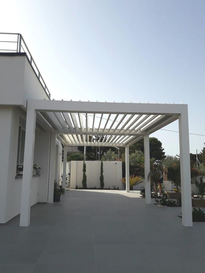 Patio moderno con pergola bianca, pavimento in cemento e vegetazione circostante sotto un cielo limpido.