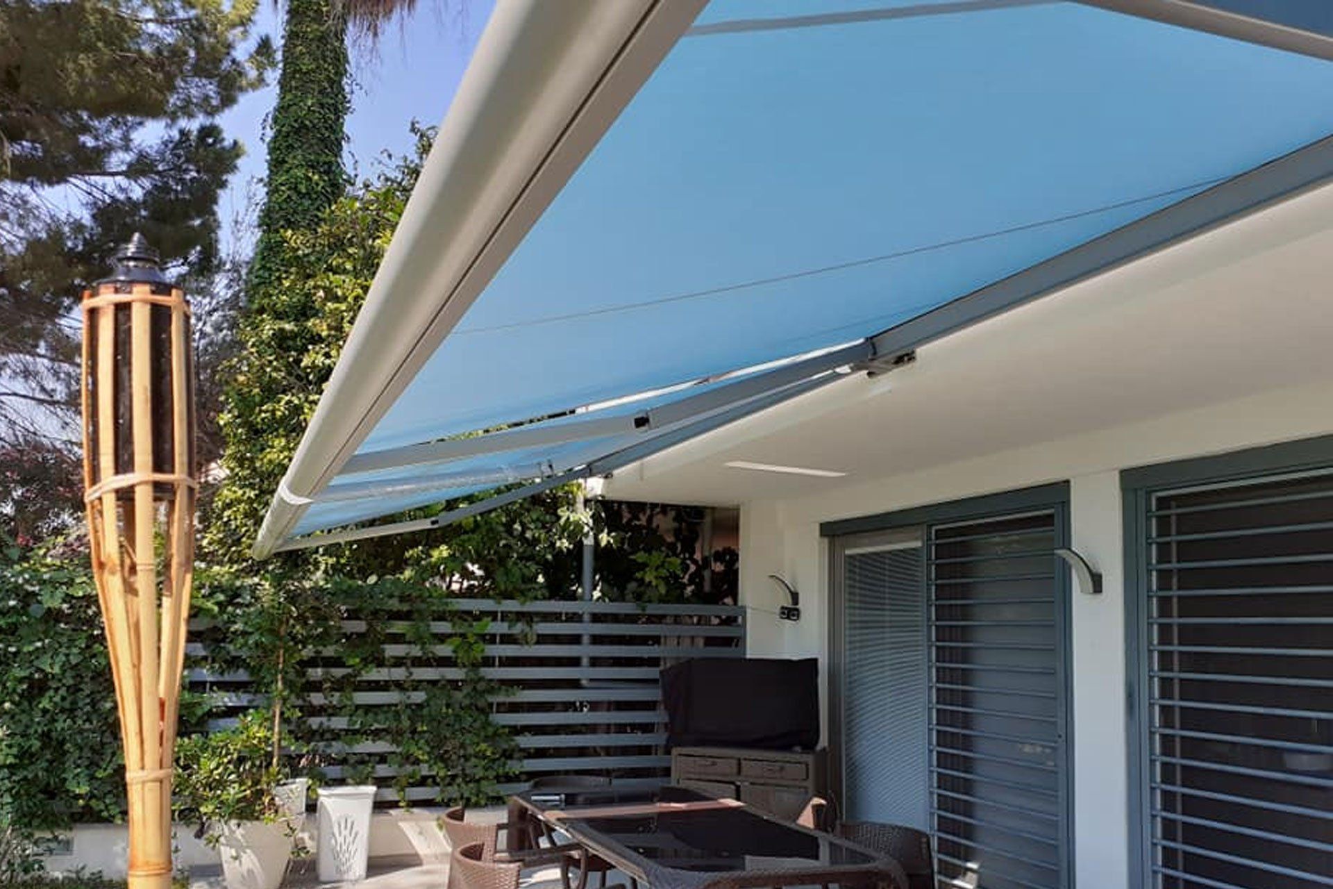 Patio con tenda da sole blu, tavolo da pranzo all'aperto e torce tiki in legno accanto a una casa bianca.